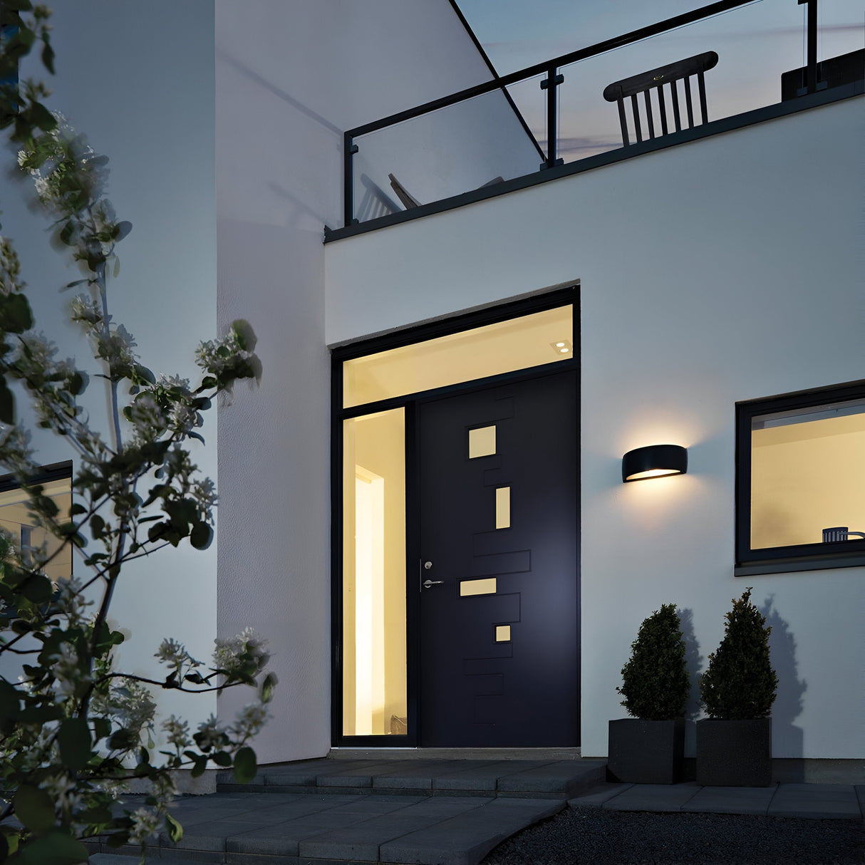 In the modern house exterior at twilight, a sleek black door with rectangular windows stands out. Warm indoor light glows through the die-cast aluminum door and surrounding windows. Enhancing the contemporary look are potted plants and a Vasa Outdoor 1 Light Wall Light in Graphite, which is IP65 rated for outdoor use.