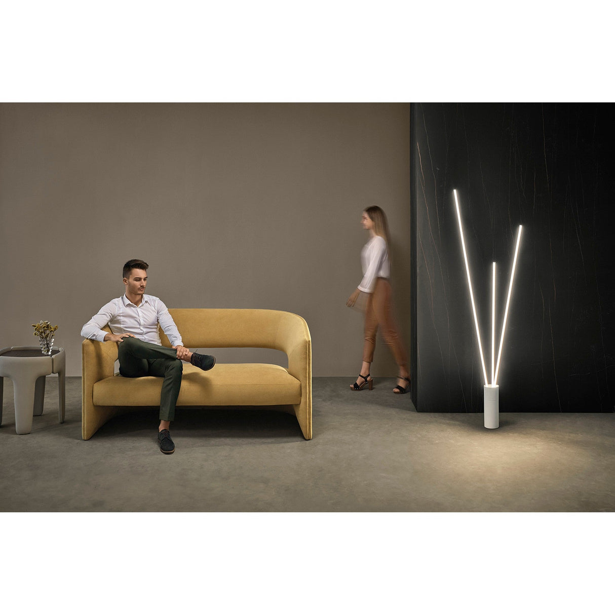 A man sits on a mustard-yellow sofa with one leg crossed, while a woman walks past a black wall. The modern room features minimal decor, a small side table, and the Vertical Dimmable LED Floor Lamp 60W 3000K - White.