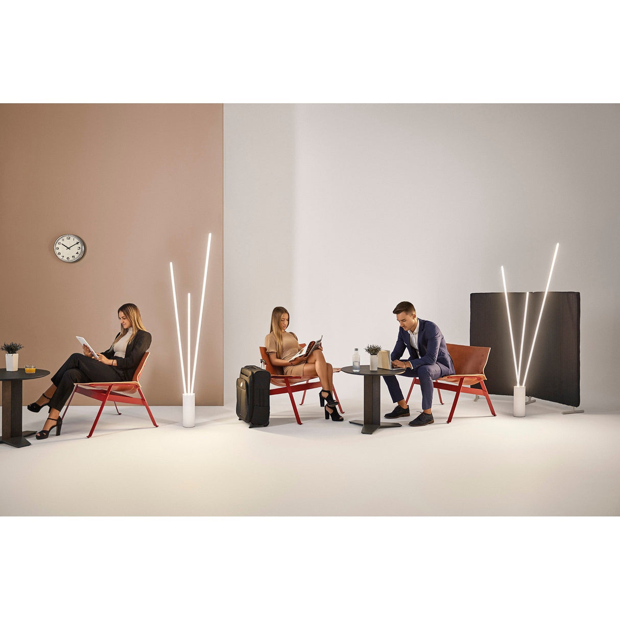 Three people sit apart in a minimalistic room on red chairs with small tables, reading or writing. A Vertical Dimmable LED Floor Lamp 60W 3000K - White and a wall clock enhance the modern style of the space.