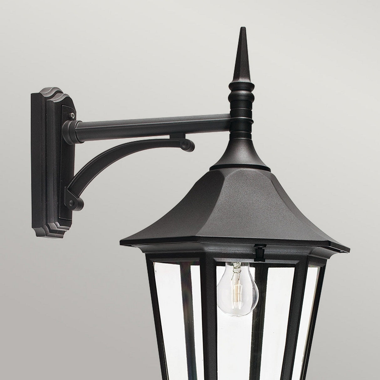 The Valencia Outdoor Large 1 Light Down Lantern in black showcases a classic die-cast aluminum design with a pointed top, elegantly mounted on an ornate black arm. This weather-resistant lantern features clear glass panels that reveal a visible light bulb inside, highlighted against a plain gray background to enhance its timeless outdoor lighting appeal.