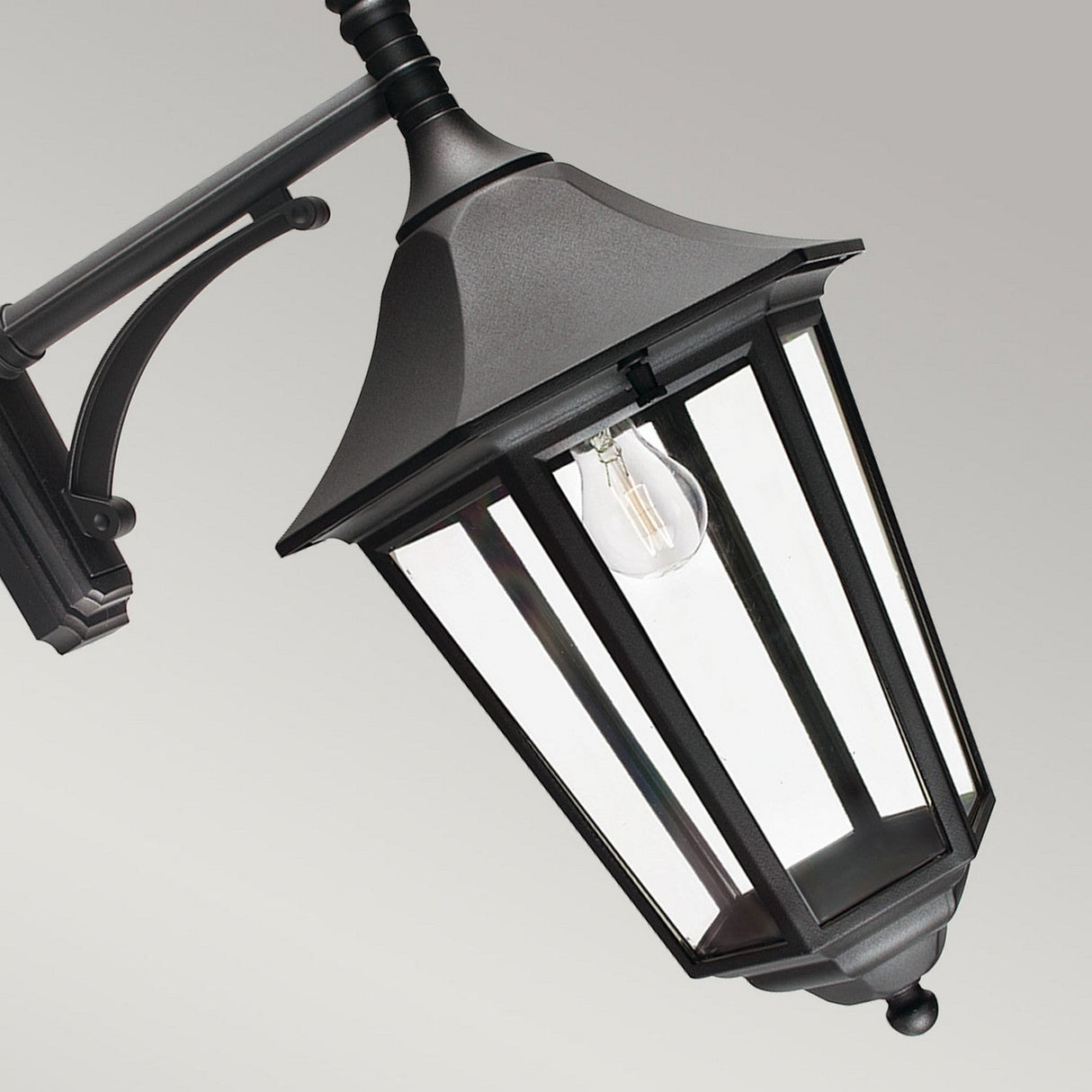 The Valencia Outdoor Large 1 Light Down Lantern - Black is an elegant lantern crafted from die-cast aluminum in a classic style. It features clear glass panels and a single exposed bulb, embodying a timeless appeal. This weather-resistant outdoor lighting fixture is securely mounted on a curved metal bracket set against a neutral gray background.