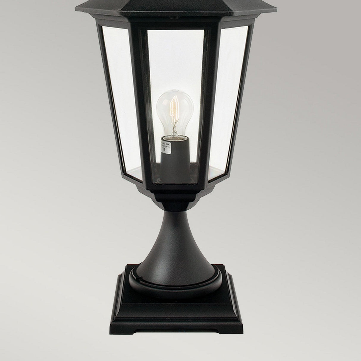 The Valencia Large 1 Light Pedestal - Black is an elegant outdoor lantern showcasing a hexagonal glass casing with a visible light bulb. Crafted from durable die-cast aluminum, this stylish lighting fixture is mounted on a square base and set against a plain gray background.