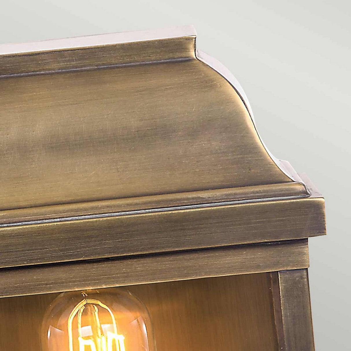 Close-up of the Victoria 1 Light Outdoor Wall Light in aged brass, showcasing its rectangular design and smooth metallic finish with a partially visible top edge. The visible filament bulb adds to its charm against a muted gray background, enhancing the lamp's timeless elegance.