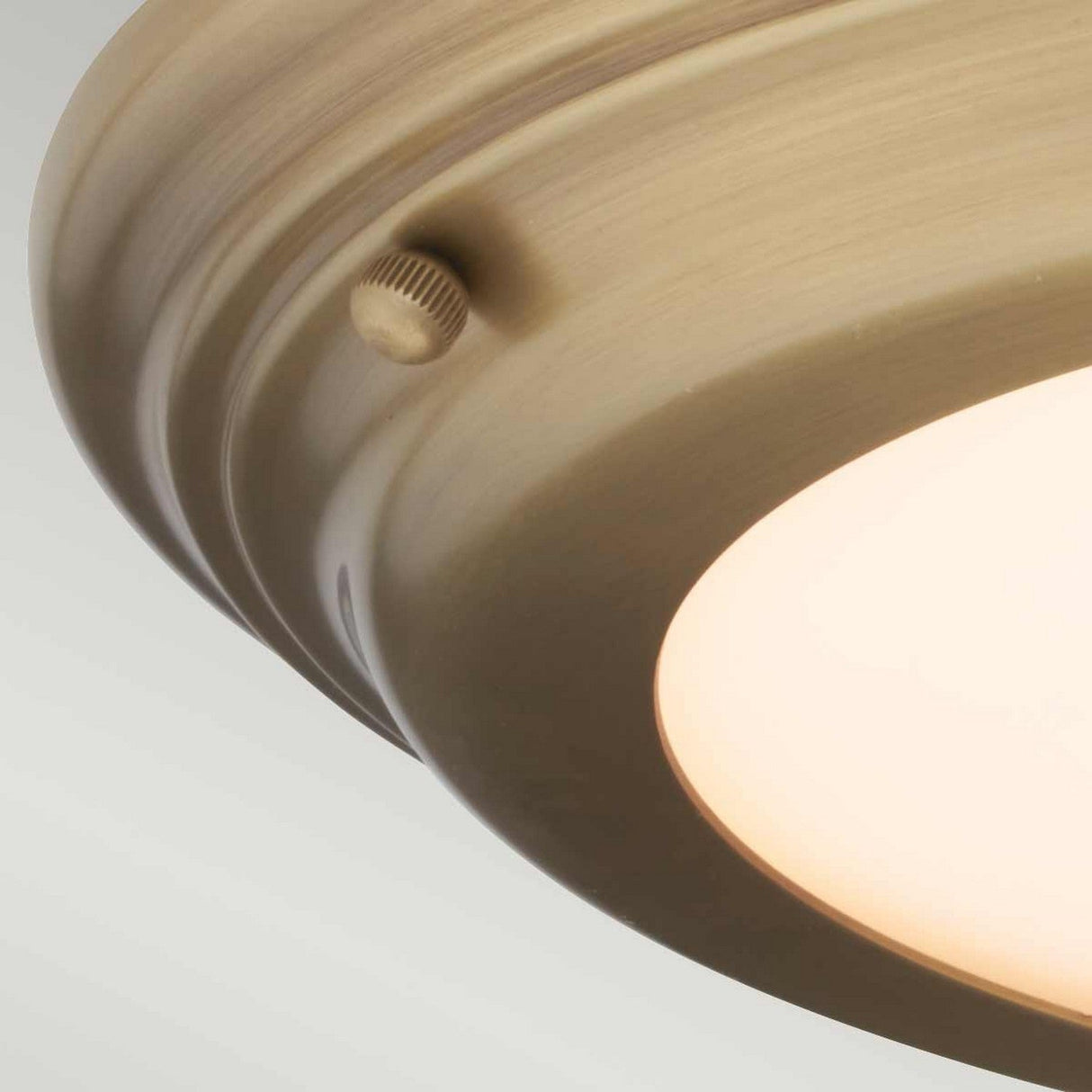 Close-up of the Welland LED Light Flush in aged brass, featuring a frosted glass cover. A small, round knob is visible on the side, set against a light gray background. This fixture elegantly combines style with modern efficiency.