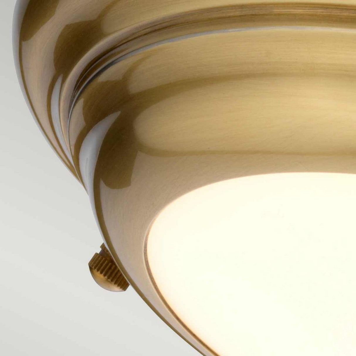 A close-up view of the Welland LED Light Mini Flush in aged brass showcases its frosted white glass shade. This fixture, designed with a mini flush look, includes a decorative side screw for securing the glass. It emits soft lighting that creates a warm glow, enhancing the rich tones of its brass finish.