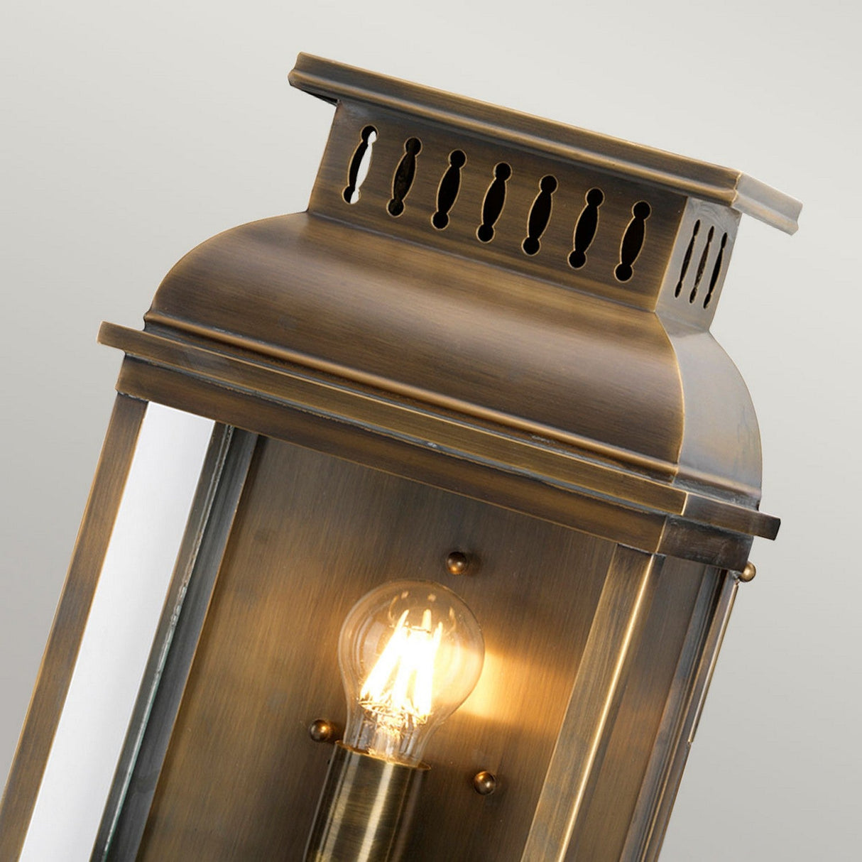 Close-up of the Westminster 1 Light Outdoor Wall Light - Aged Brass, featuring a radiant bulb within its antique bronze finish. This outdoor wall fixture boasts a rectangular design with a decorative top vent and clear glass panels, emanating an aged brass allure against a soft, neutral light gray background.