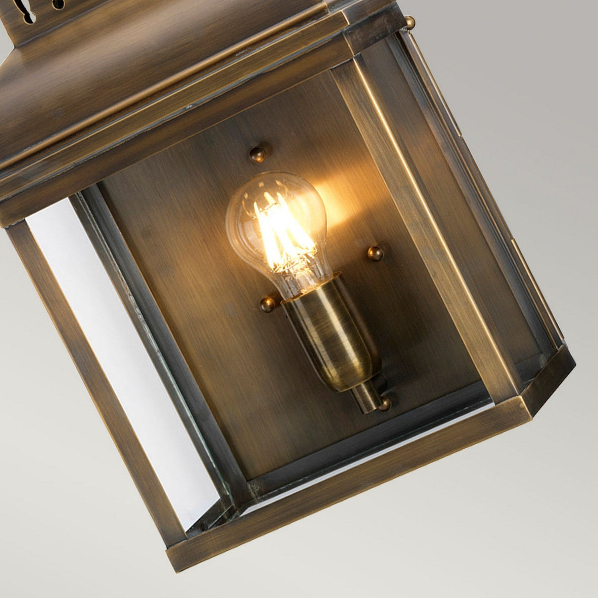 A close-up of the Westminster 1 Light Outdoor Wall Light - Aged Brass showcases its glowing bulb inside. This fixture features an aged brass finish and transparent glass panes, casting a warm glow in a classic design.
