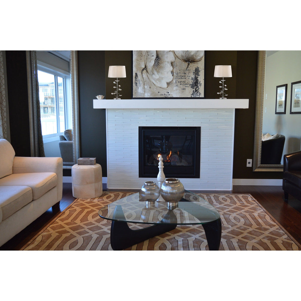 A modern living room features a white brick fireplace with abstract art and Willow 1 Light Crystal Table Lamps—chrome bases with white shades—on the mantel. A glass coffee table with crystal accents sits on a geometric rug between beige and black chairs. Large mirrors flank the fireplace.