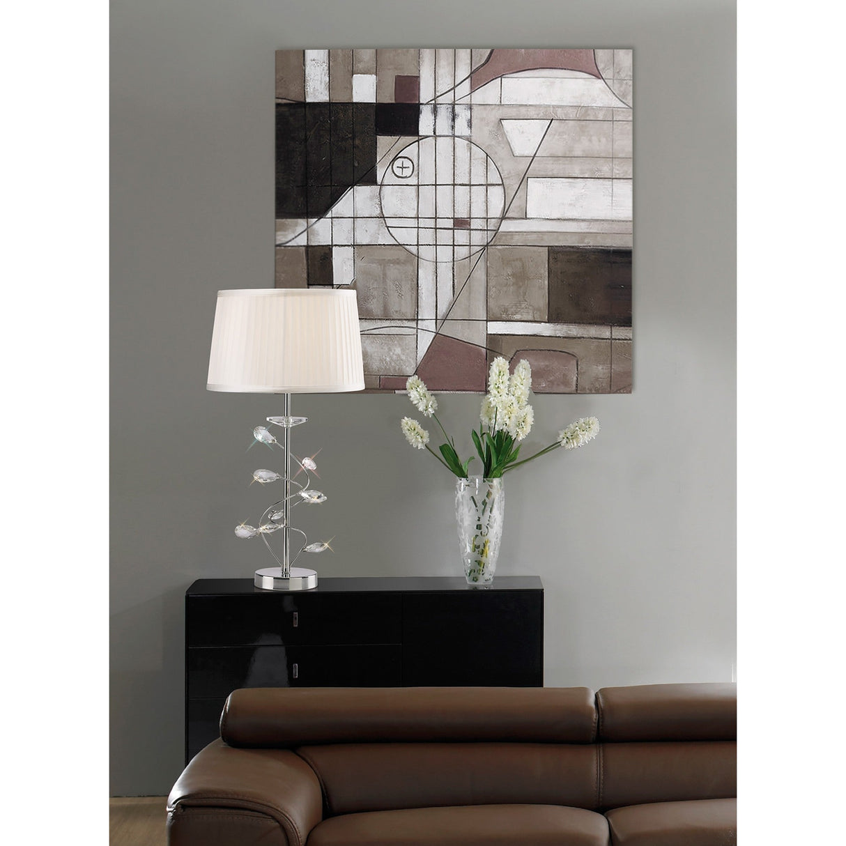 A modern living room includes a brown leather sofa, a black sideboard with a glass vase of white flowers, and the Willow 1 Light Crystal Table Lamp—chrome finish with white shade—beside an abstract geometric painting on a gray wall.