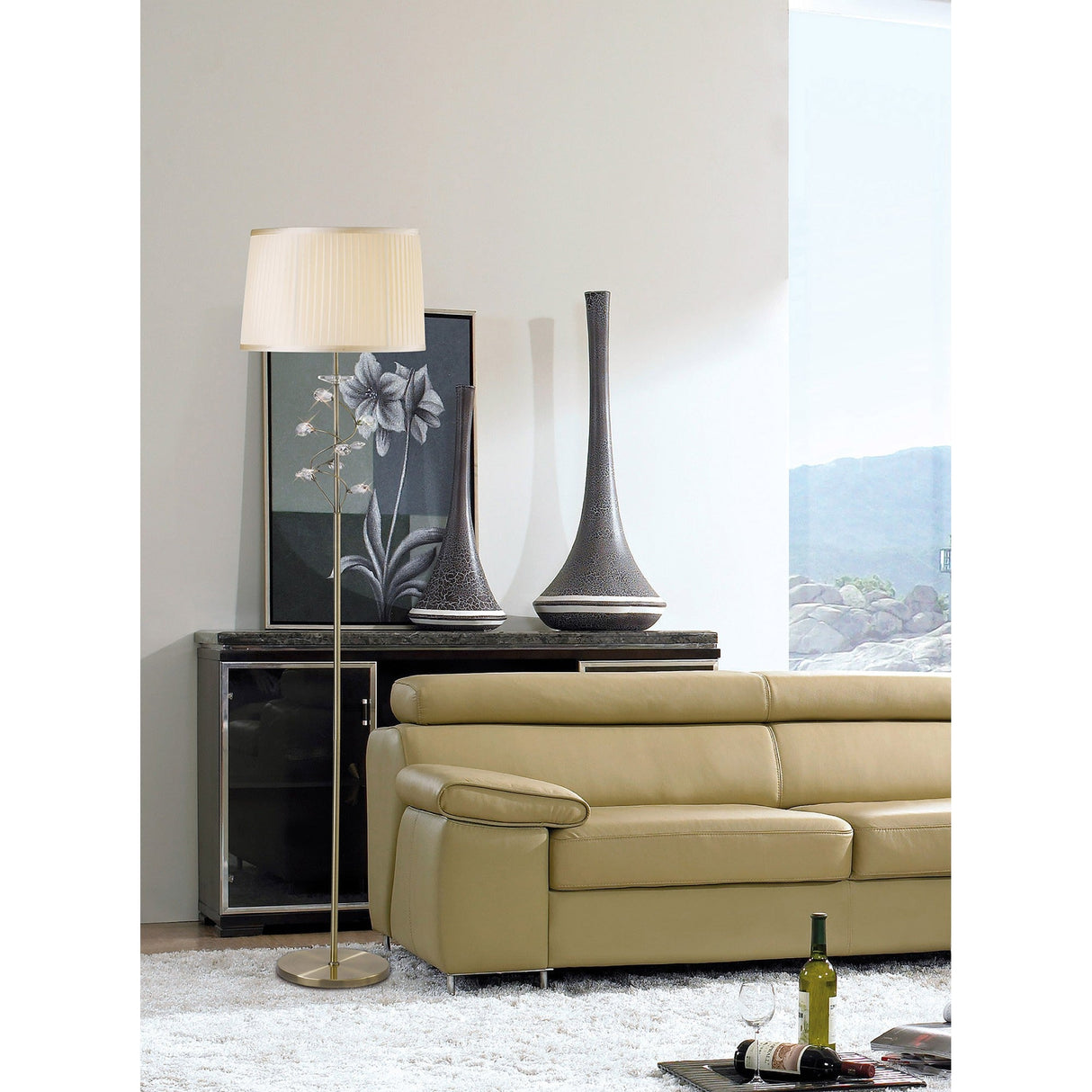 A modern living room with a beige leather sofa, the Willow 1 Light Crystal Floor Lamp in brass with a cream shade, two tall vases, framed floral art, a glass cabinet, and a coffee table displaying an open magazine and wine bottle.