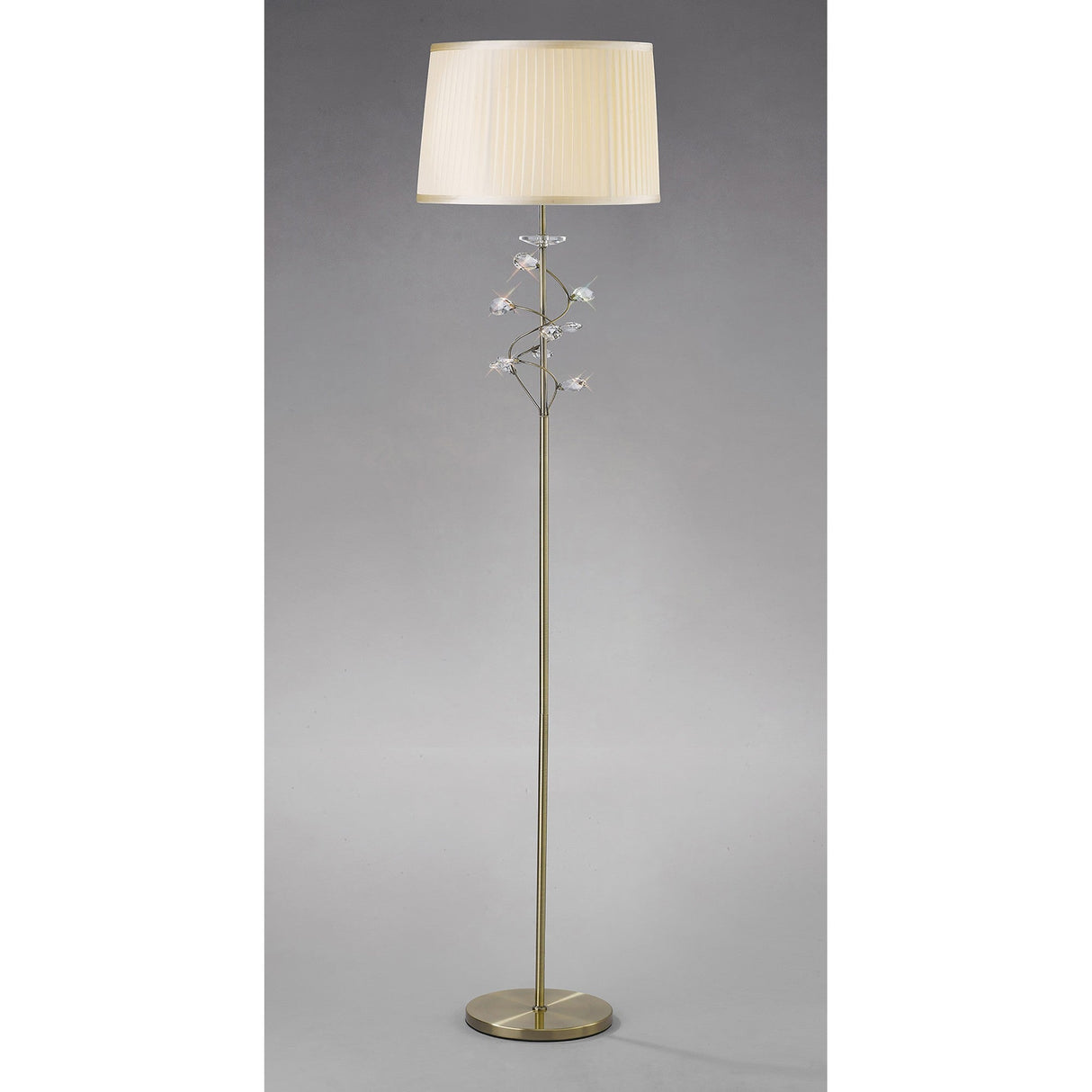 The Willow 1 Light Crystal Floor Lamp features a round base, slender antique brass pole, cream pleated shade, and decorative crystal flower accents on the stem, all set against a plain light gray background.