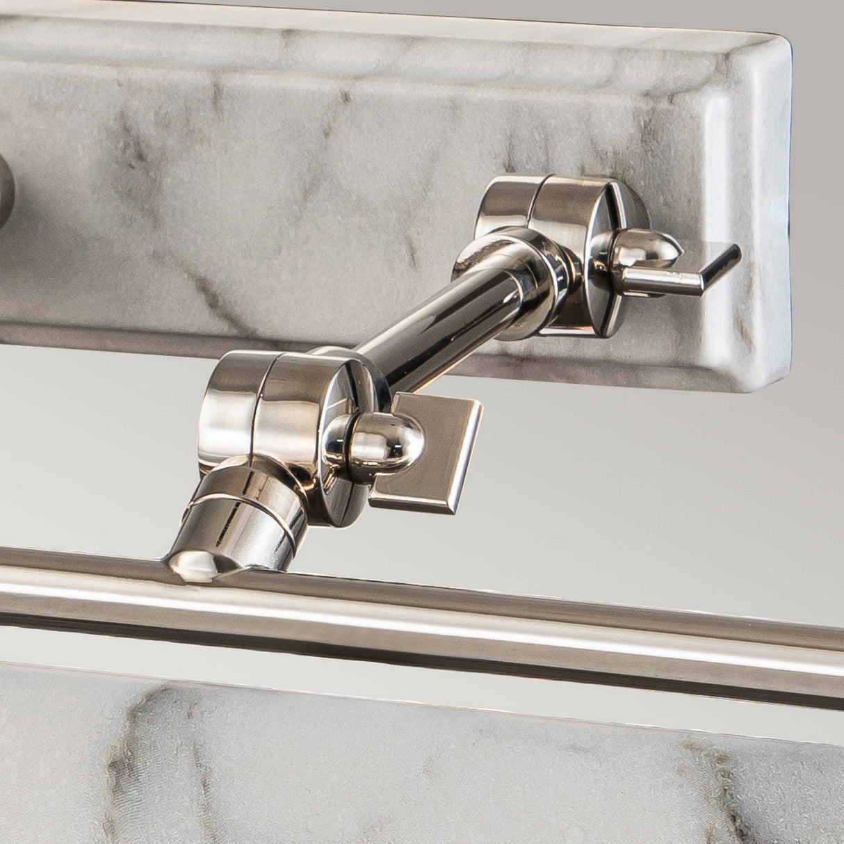 A close-up of the Winchfield 4 Light Large Picture Light showcases its wall-mounted metal bracket holding a horizontal rod, set against a white marble effect background. The bracket is finished in polished nickel with adjustable components, making it perfect for illuminating artwork.