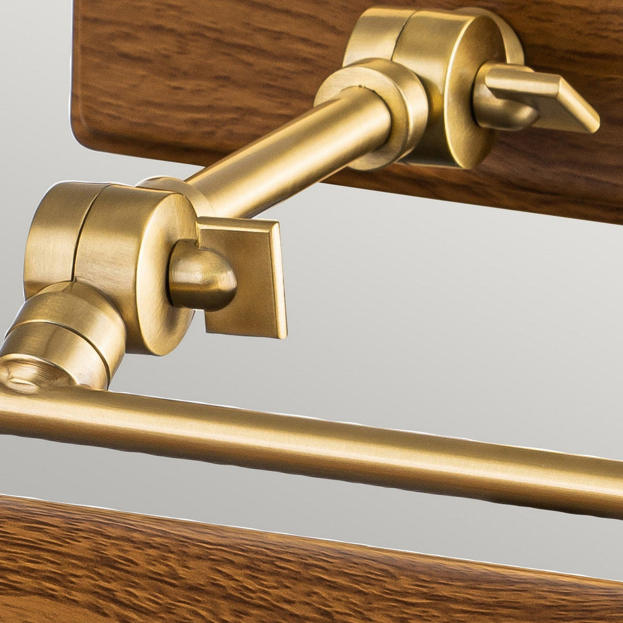 Close-up of the Winchfield 2 Light Medium Picture Light in aged brass attached to a piece of oak-effect wood. The intricate design showcases rods and connection points, highlighting craftsmanship and functionality. The wood, reminiscent of Mid Century Modern style, boasts a dark, polished finish.