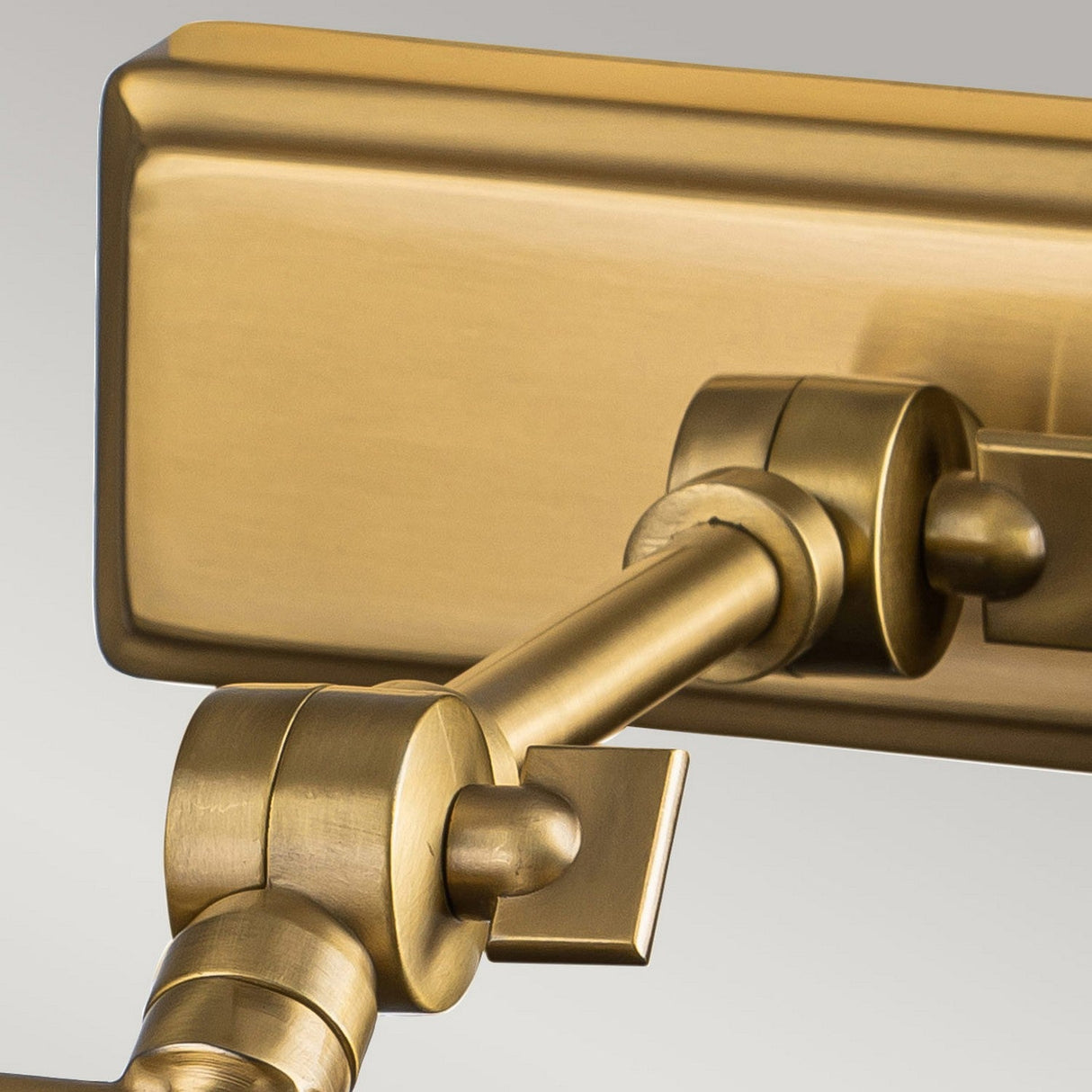 Close-up of the Winchfield 2 Light Medium Picture Light - Aged Brass, featuring an adjustable hinge mechanism with a rectangular base. Reminiscent of Mid Century Modern design, the hinge comprises cylindrical components and a connecting rod, showcasing an aged brass finish with sleek polish against a neutral background.