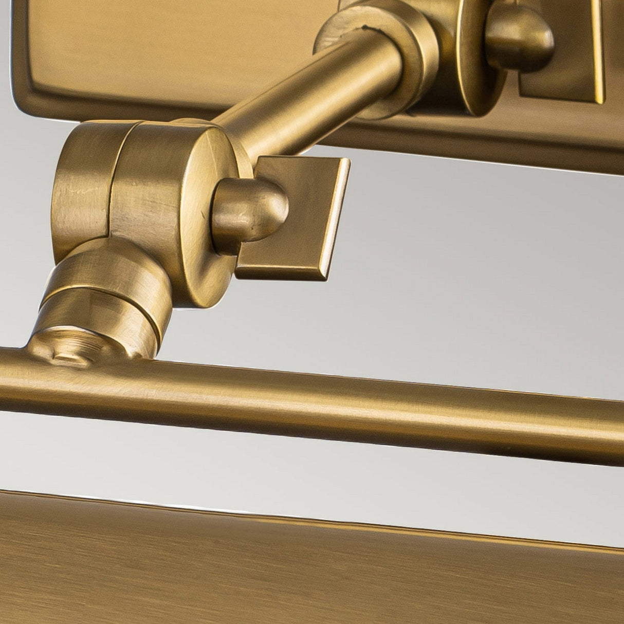 Close-up of an aged brass-colored component from the Winchfield 2 Light Medium Picture Light, attached to a metal structure. Its design reminiscent of Mid Century Modern style features a smooth, shiny finish that highlights the intricate details of its construction.