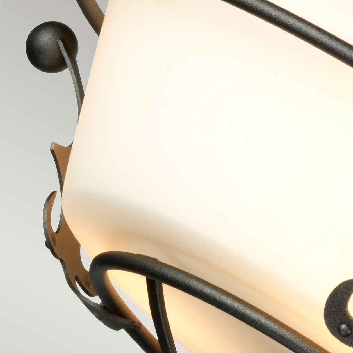 Close-up of the Windsor 3 Light Semi-Flush - Graphite decorative fixture showcasing a frosted white glass shade and black wrought iron accents. This fixture embodies a regal design with circular and curved elements, all set against a soft gray gradient, featuring an elegant graphite finish.