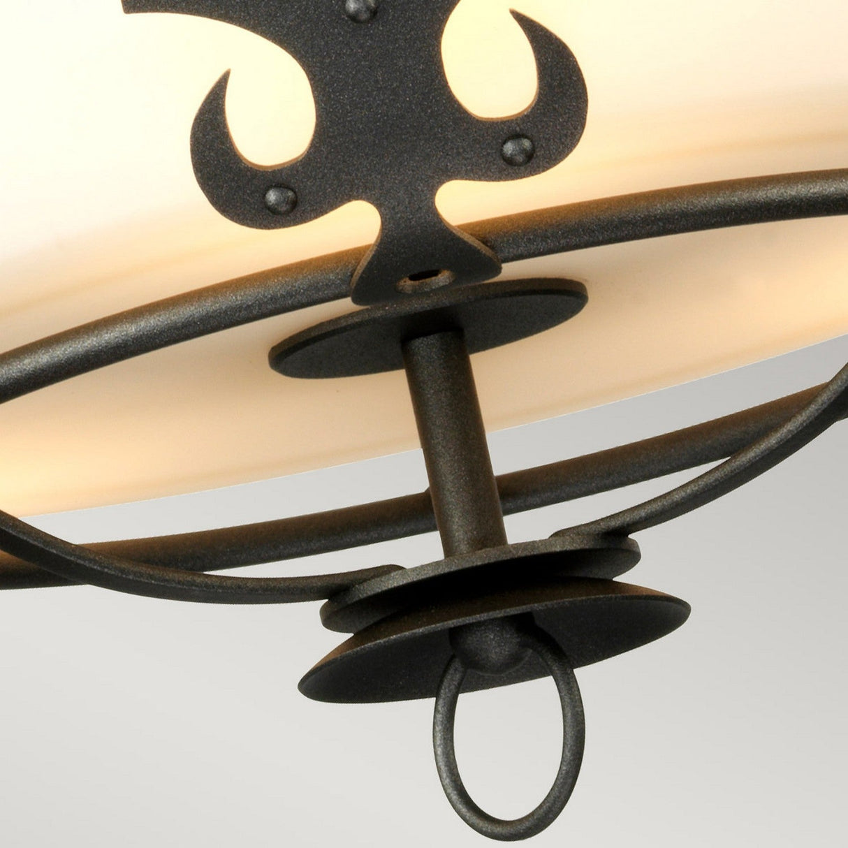 A close-up of the Windsor 3 Light Semi-Flush - Graphite chandelier showcases its cream-colored glass shade and graphite finish. The design highlights ornate, curved metalwork and sturdy fixtures, merging elegance with rustic charm against a plain, light gray background.