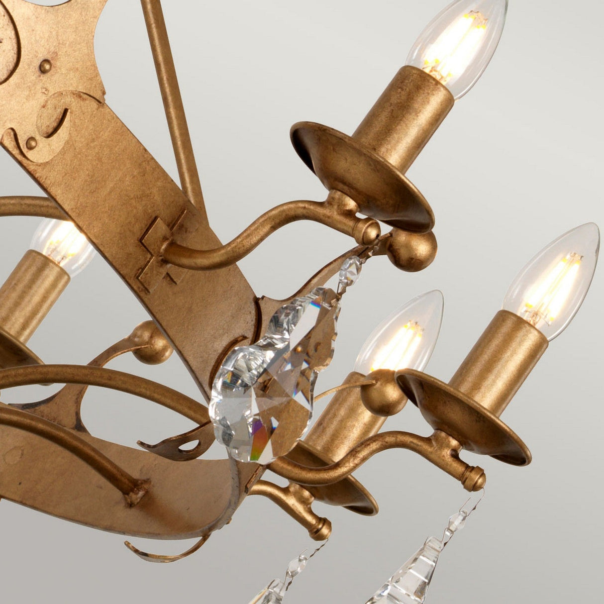 A close-up of the Windsor 12 Light Chandelier - Gold Patina highlights British craftsmanship with its intricate, aged brass frame. It boasts twelve candle-like bulbs and delicate crystal pendants that hang elegantly from its structure, accentuated by a subtle gold patina finish against a neutral backdrop.