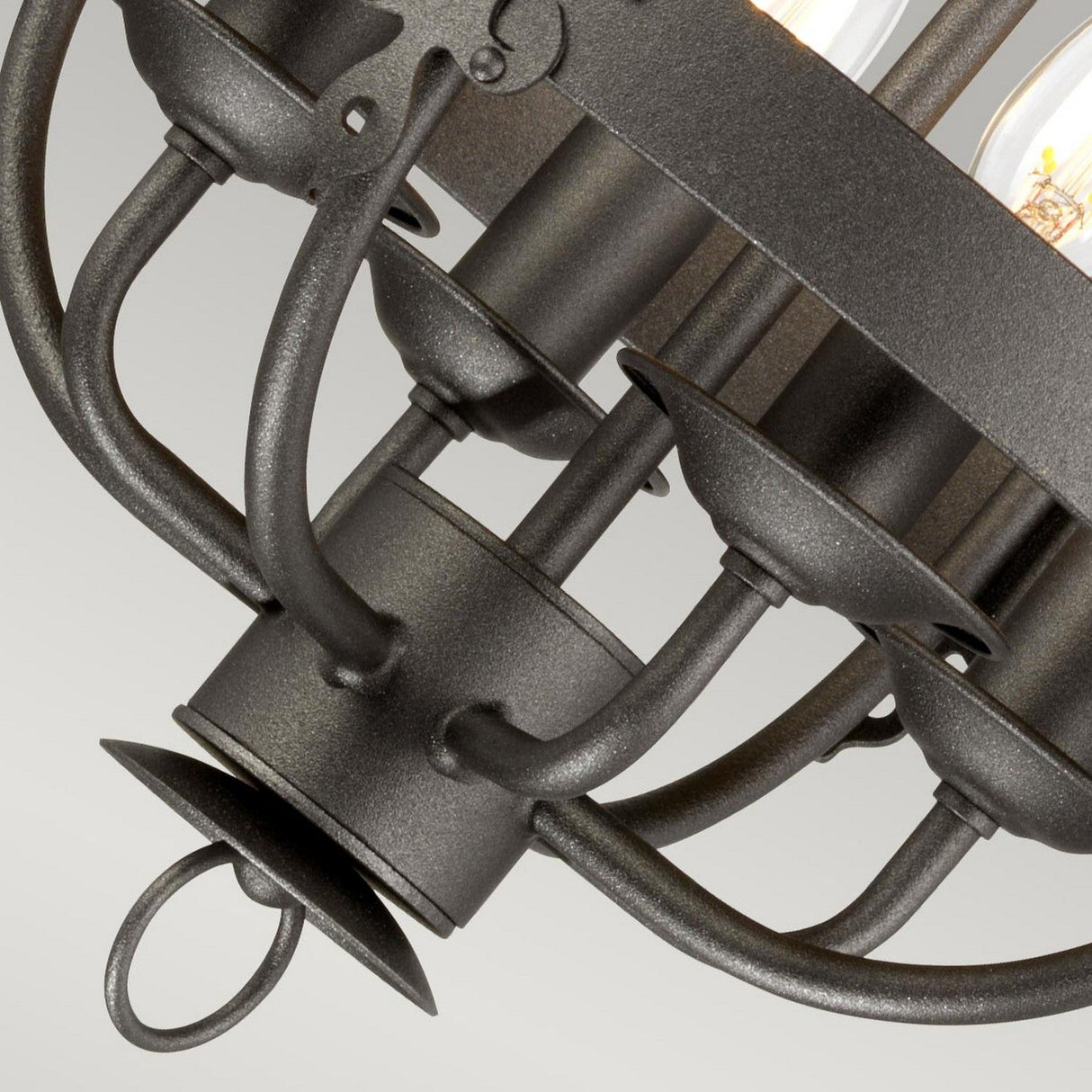 Close-up of the Windsor 4 Light Chandelier in graphite, showcasing its black metal construction with a rustic finish, hand-crafted detailing, and multiple candle-shaped light bulbs. The fixture features elegantly curved arms and a circular centerpiece, set against a plain, light gray background.