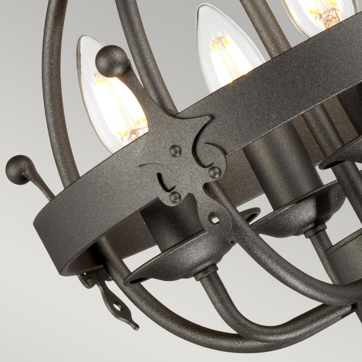 A close-up of the Windsor 4 Light Chandelier in a rustic, graphite finish showcases candle-like light bulbs. The piece is intricately designed with hand-crafted detailing and a decorative band embellished with star-shaped accents, enhancing its antique look. The chandelier's lights cast a warm, soft glow.