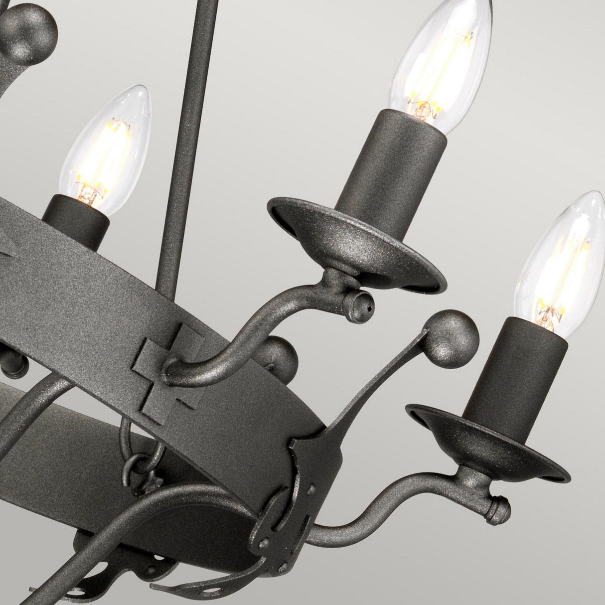 Close-up of the Windsor 6 Light Chandelier - Graphite in a dark metal with an elegant graphite finish. Featuring three lit candle-shaped bulbs, the design showcases curved arms and a circular frame adorned with decorative crosses. The plain light gray backdrop enhances its sophisticated lighting appeal.