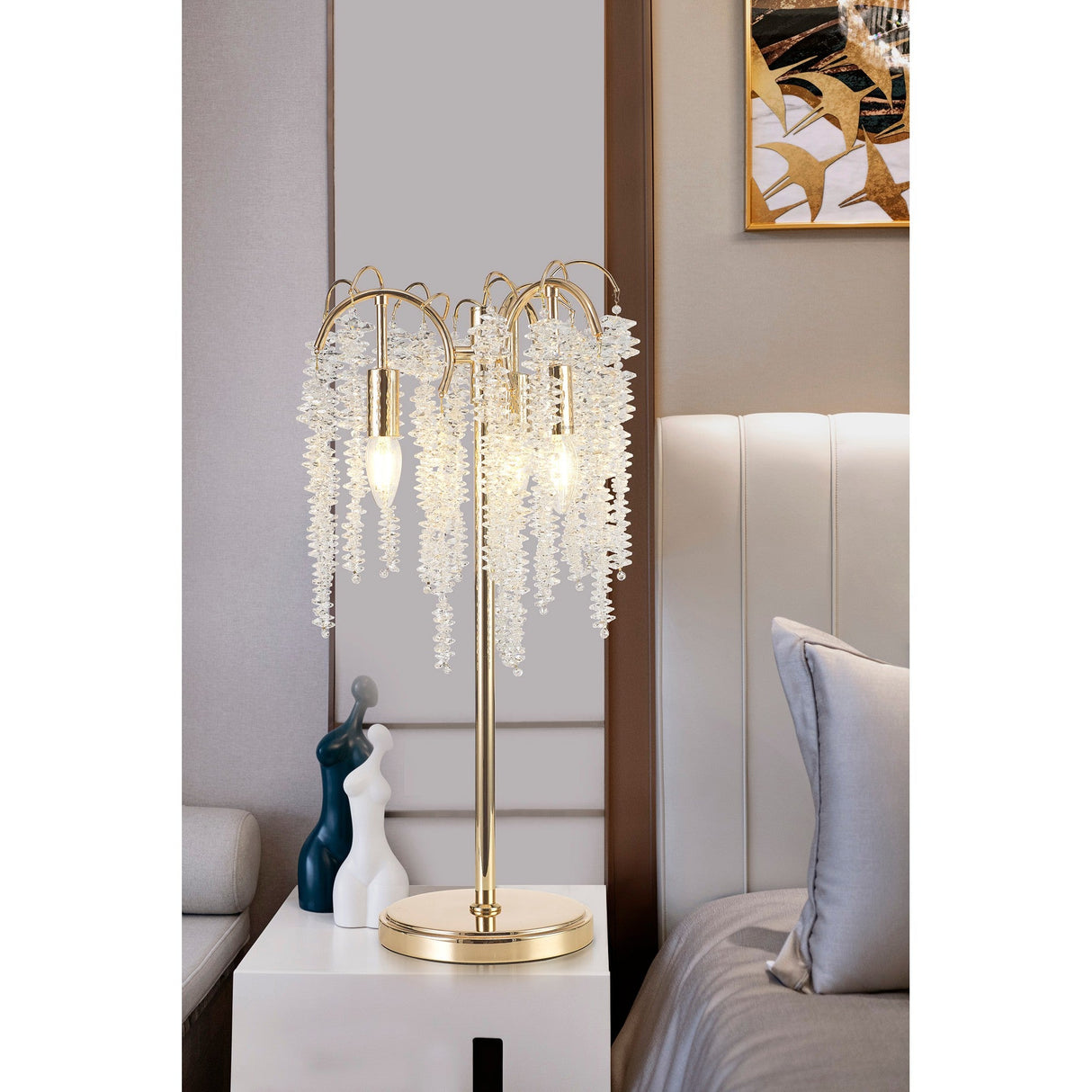 The Wisteria 3 Light Table Lamp in gold with clear crystal clusters provides dimmable lighting on a bedside table beside a modern bed with a white headboard and gray pillow in a stylish bedroom.