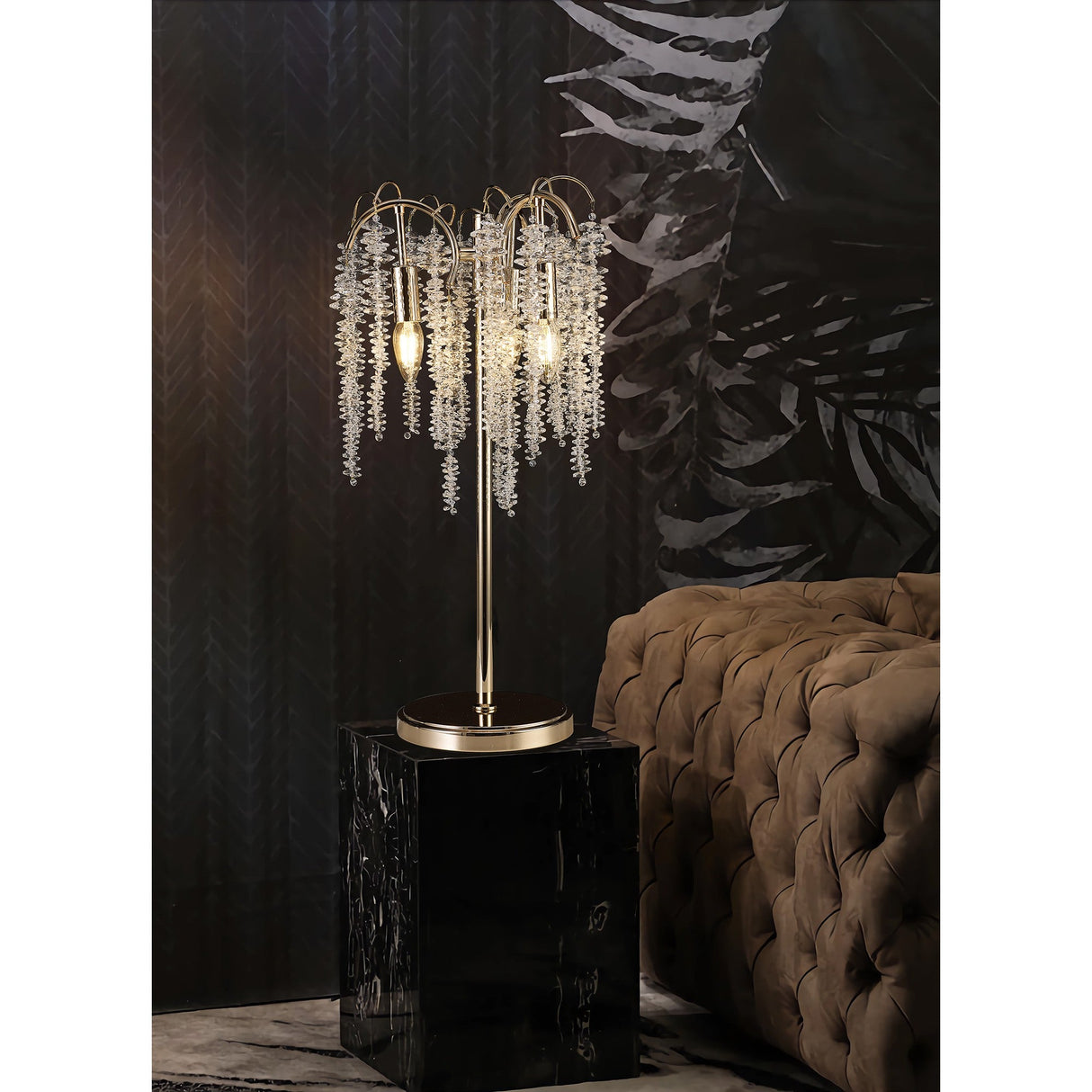 The Wisteria 3 Light Table Lamp in gold with clear crystal clusters sits on a black marble pedestal beside a tufted brown velvet sofa, casting elegant dimmable lighting against a dark, leaf-patterned wall.