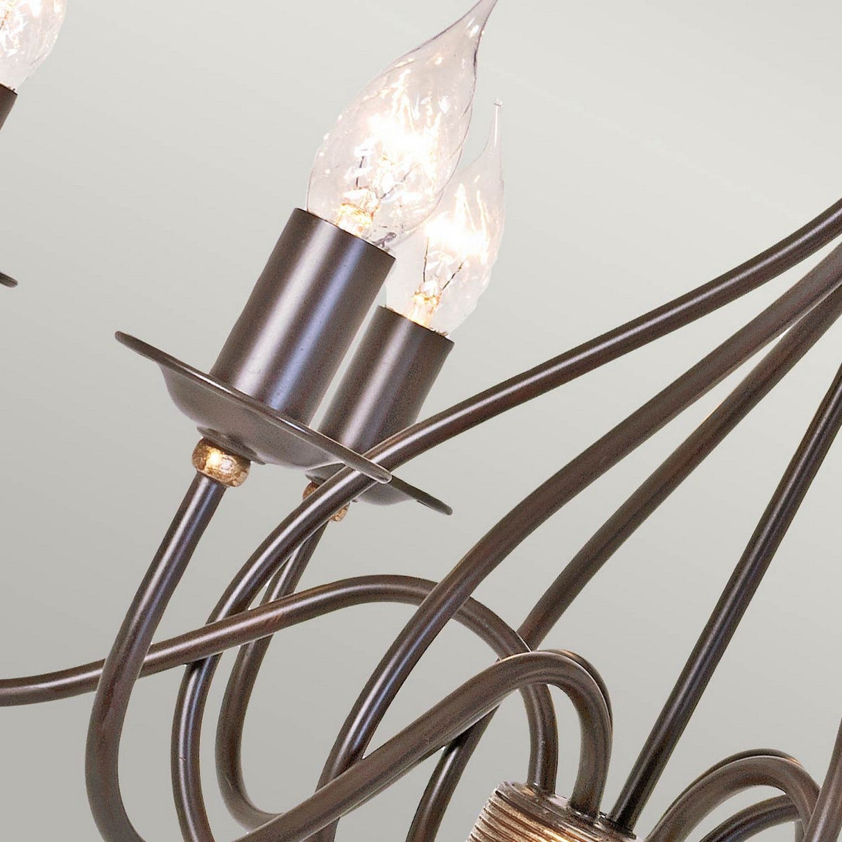 A close-up of the Windermere 6 Light Chandelier in rust and gold shows its sleek, modern design featuring curving wrought iron rods. The handcrafted piece is adorned with metallic arms and candle-shaped LED bulbs, all set against a light-colored background.