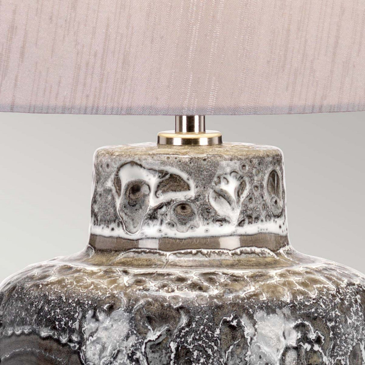 A close-up of the Woolwich 1 Light Table Lamp reveals its textured ceramic base in a graphite hue, showcasing a gray and white marbled effect with a smooth, glossy finish. The light beige fabric shade casts an ambient glow, providing a soft contrast to the intricate patterns on the base.