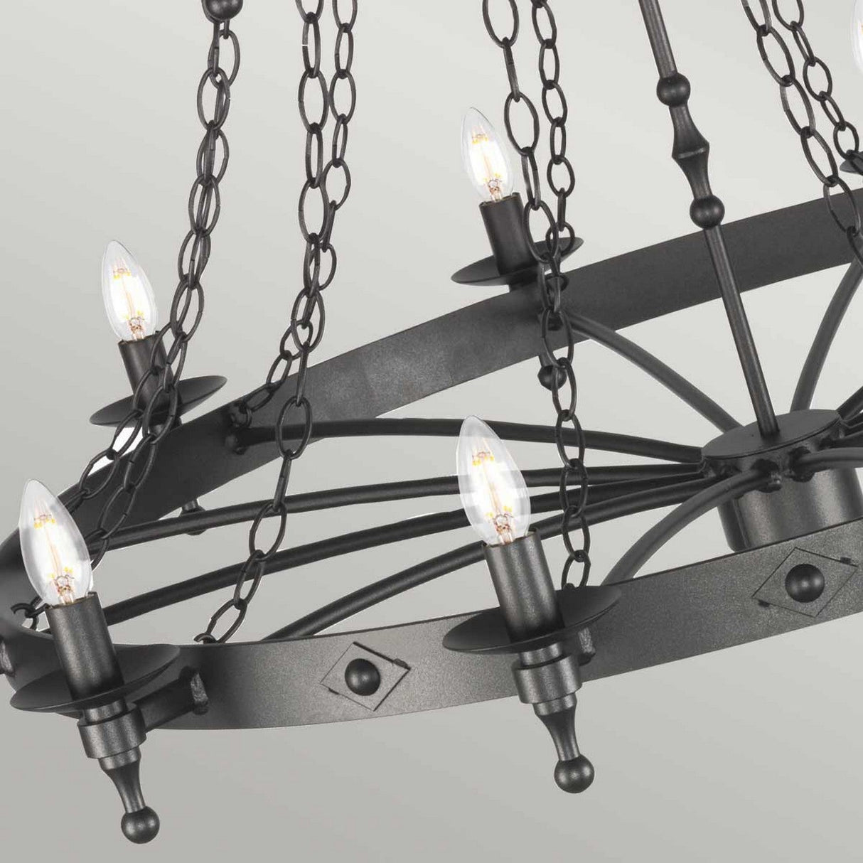 A detailed view of the Warwick 18 Light Chandelier in Graphite Black reveals its circular wrought iron construction with a series of candle-shaped light bulbs. The medieval-inspired design includes decorative chains and a geometric pattern, set against a light gray backdrop.