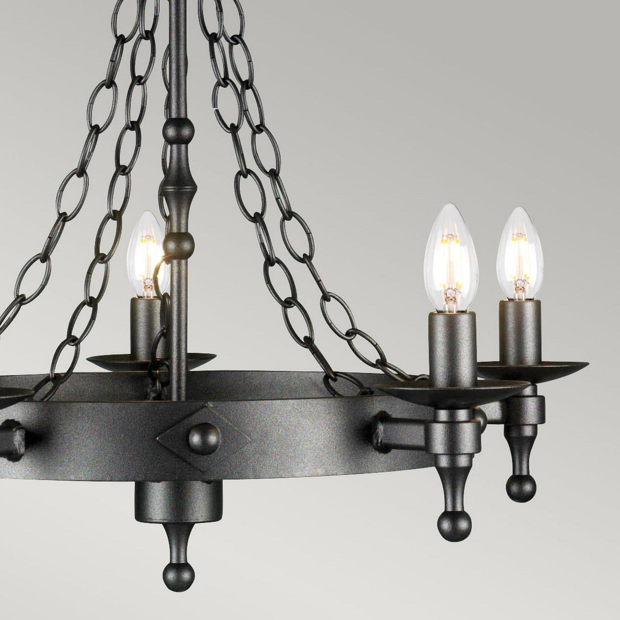 The Warwick 5 Light Chandelier in Graphite Black boasts a circular design, housing five candle-style bulbs within a frame inspired by wrought iron. Suspended by durable chains, it combines modern elegance with rustic charm, reminiscent of medieval lighting against a neutral gray backdrop.