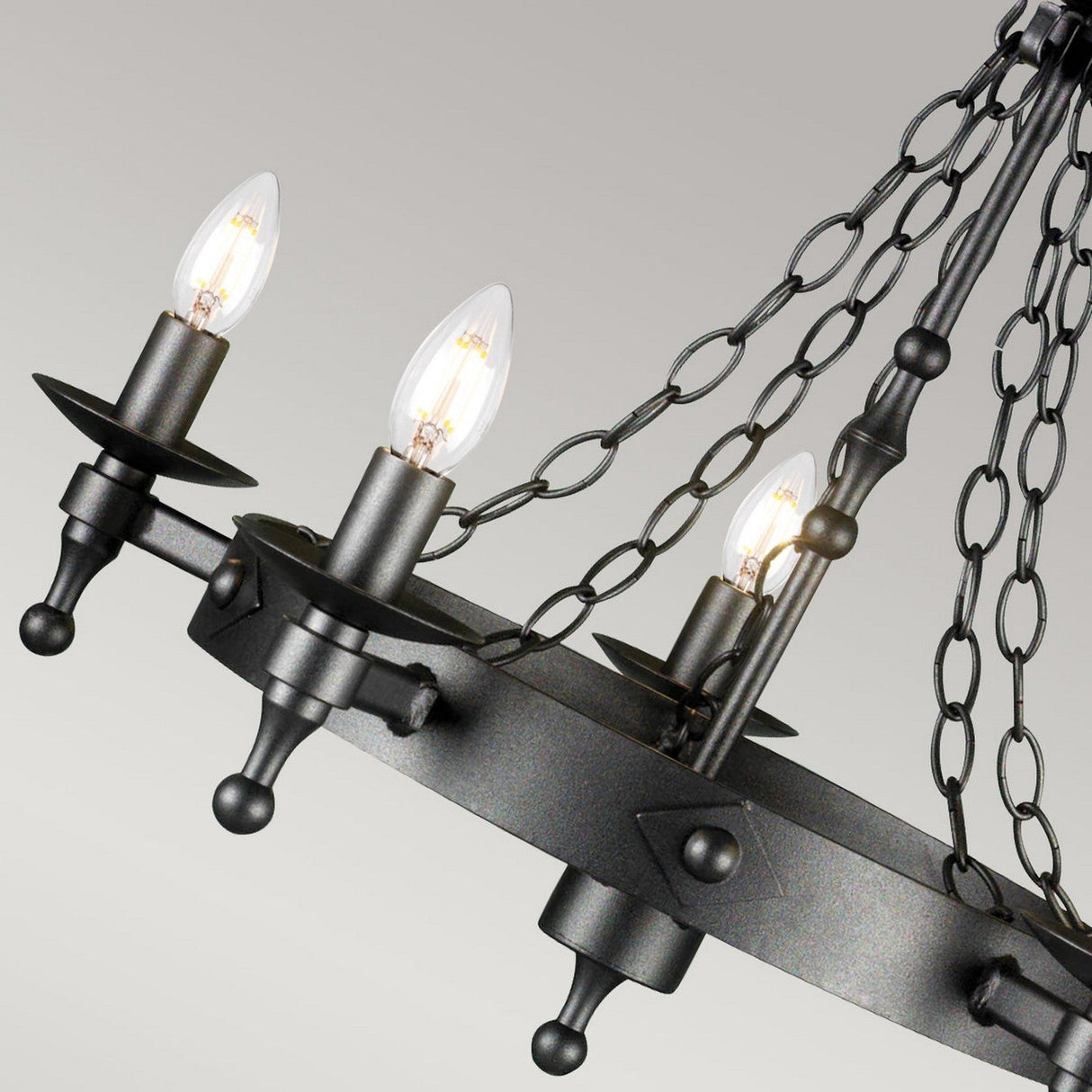 A close-up of the Warwick 5 Light Chandelier in Graphite Black, featuring a rustic, wrought iron-inspired frame and five candle-shaped light bulbs. This medieval-style chandelier showcases a round frame with chains connecting to the center, set against a plain background.