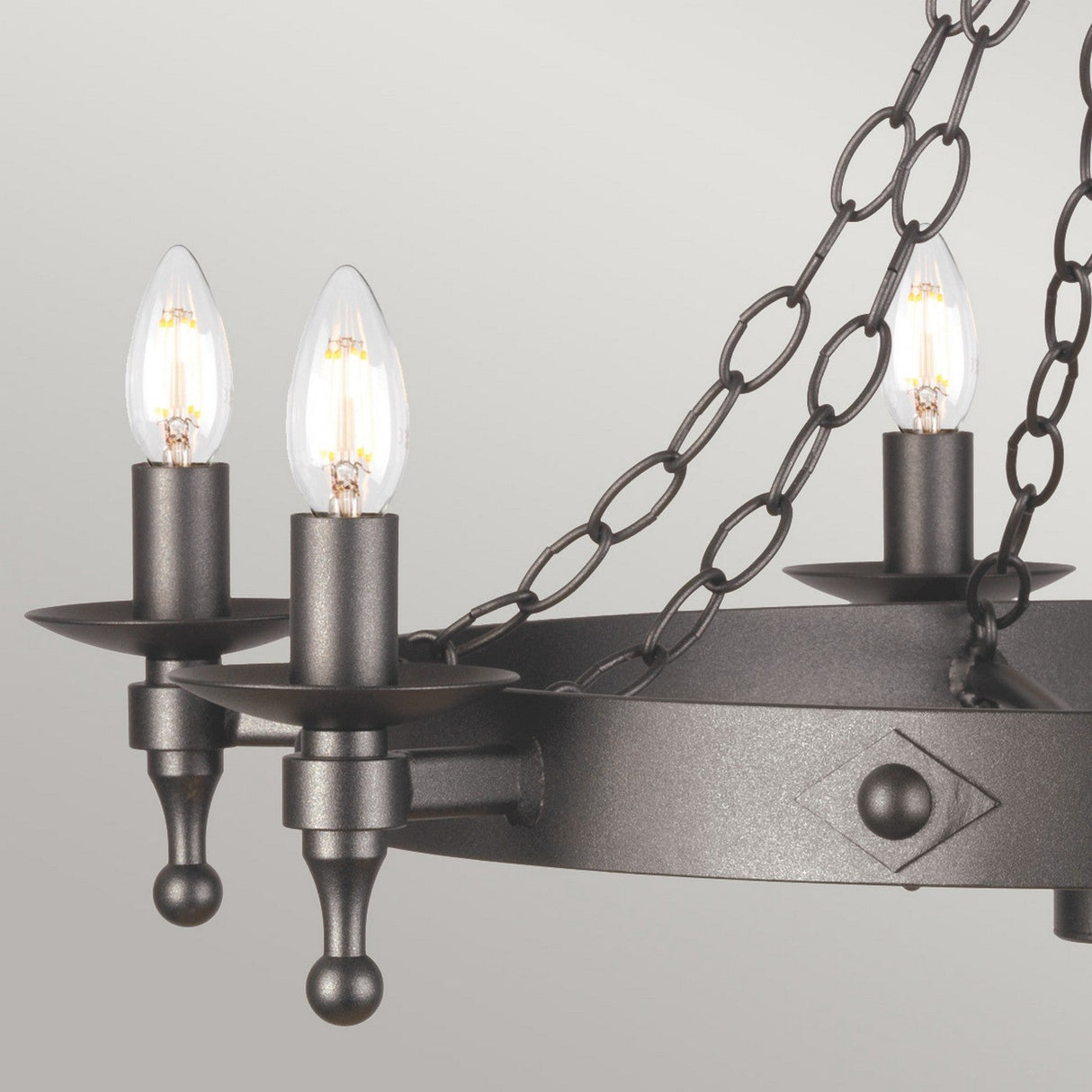 Close-up of the Warwick 6 Light Chandelier in Graphite Black, showcasing its ornate design with six candle-shaped bulbs on a circular wrought iron frame. The fixture is adorned with decorative chains and has a matte, dark finish against a neutral background.
