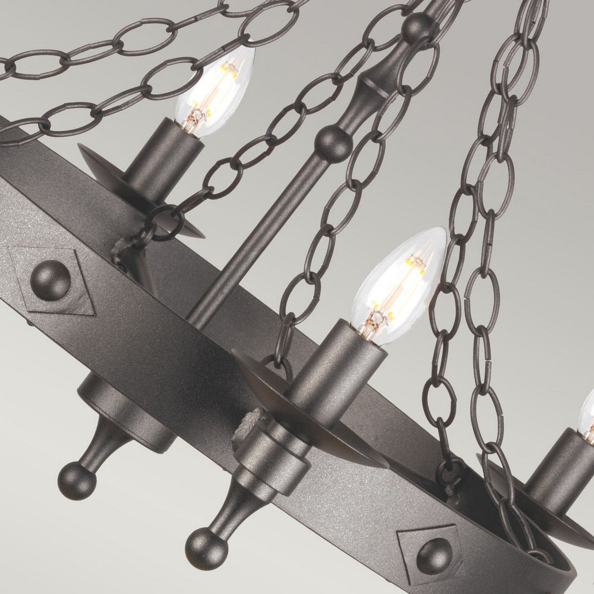Close-up of the Warwick 6 Light Chandelier in Graphite Black, crafted from black wrought iron and featuring a circular design with chain links. It boasts candle-like light bulbs and decorative knobs, evoking a vintage look. The light background enhances the fixture's ornate details.
