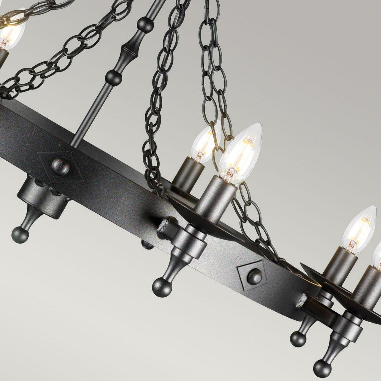 A close-up of the Warwick 8 Light Chandelier in Graphite Black reveals its rustic wrought iron design with candle-shaped bulbs, exuding an air of sophisticated charm. It hangs elegantly from chains, featuring a circular frame adorned with decorative elements, while the soft gray background enhances its timeless allure.