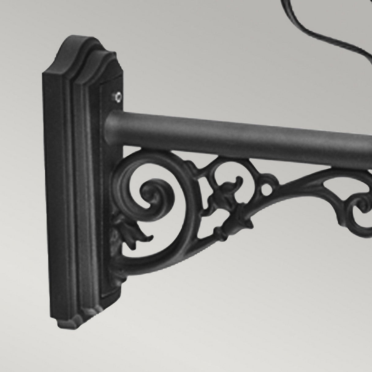 The Wilmslow 1 Light Outdoor Wall Light - Black is an elegant black wrought iron fixture featuring intricate scrollwork, evoking the charm of Victorian street lamps. It is designed to hold a hanging sign and is crafted to withstand various weather conditions when mounted on a flat, grey wall.