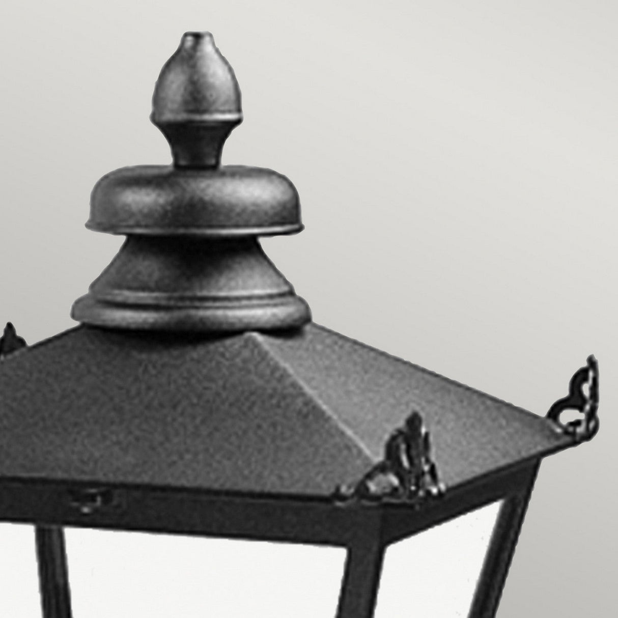 Close-up of the Wilmslow 1 Light Outdoor Wall Light in black featuring a weather-resistant design, vintage-style with a decorative metal top and glass panels, reminiscent of Victorian street lamps, set against a plain gray background.