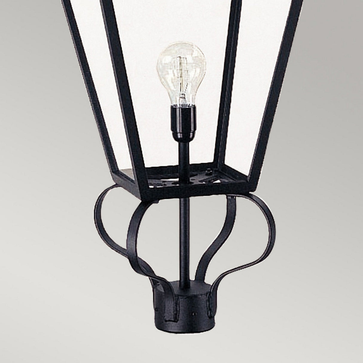 The Wilmslow 1 Light Outdoor (Head Only) - Black offers a close-up view of its Victorian-inspired design, ideal for enhancing garden illumination. It features a clear glass bulb encased within the black metal lantern head, complete with a slightly flared top and decorative curved metal elements at the base. This piece stands out elegantly against a plain gray background, making it an excellent choice for sophisticated outdoor lighting.