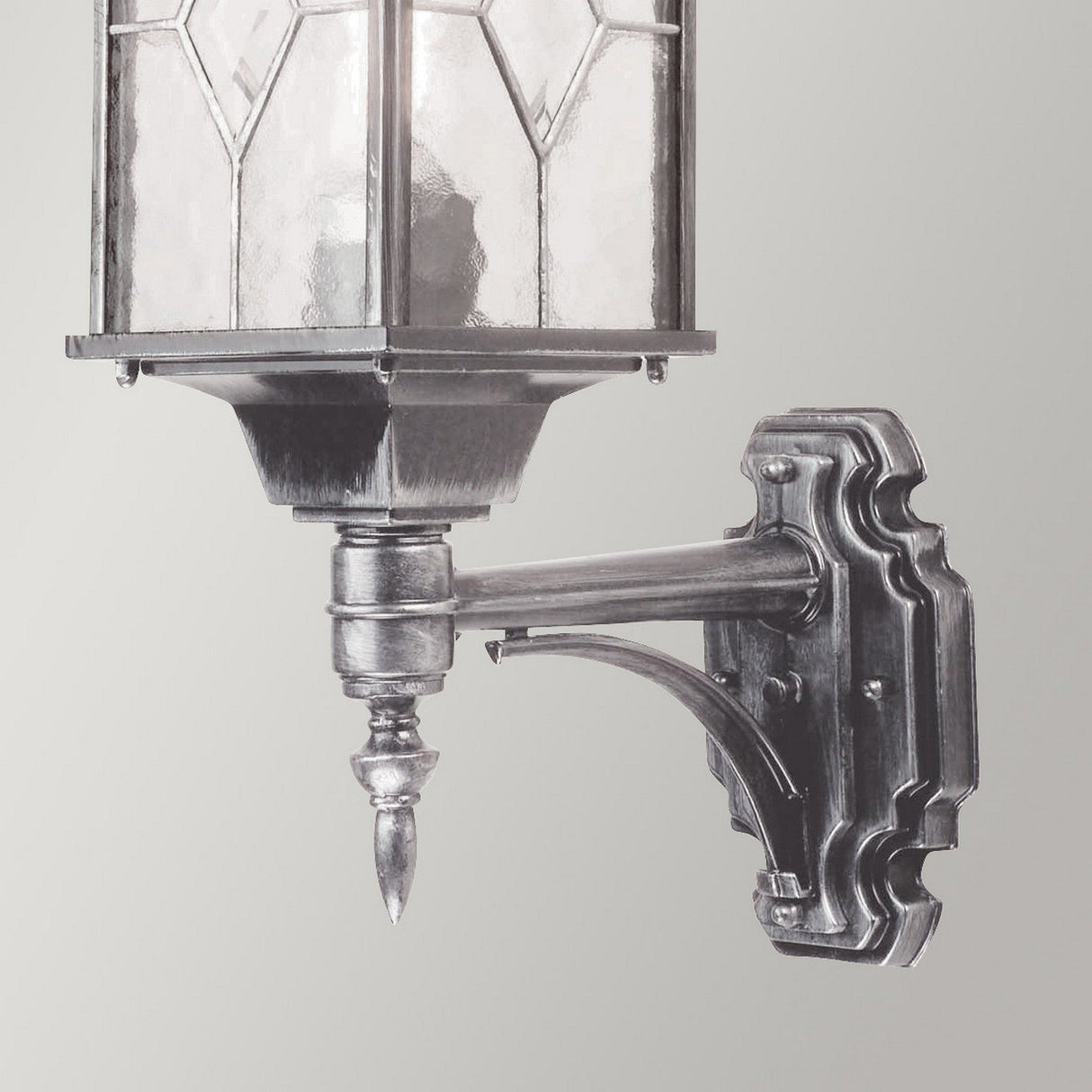 The Wexford 1 Light Outdoor Up Wall Light in Black/Silver showcases a traditional lantern design with a vintage-style metal frame and leaded glass panels, gracefully mounted to a decorative metal bracket against a light gray background.