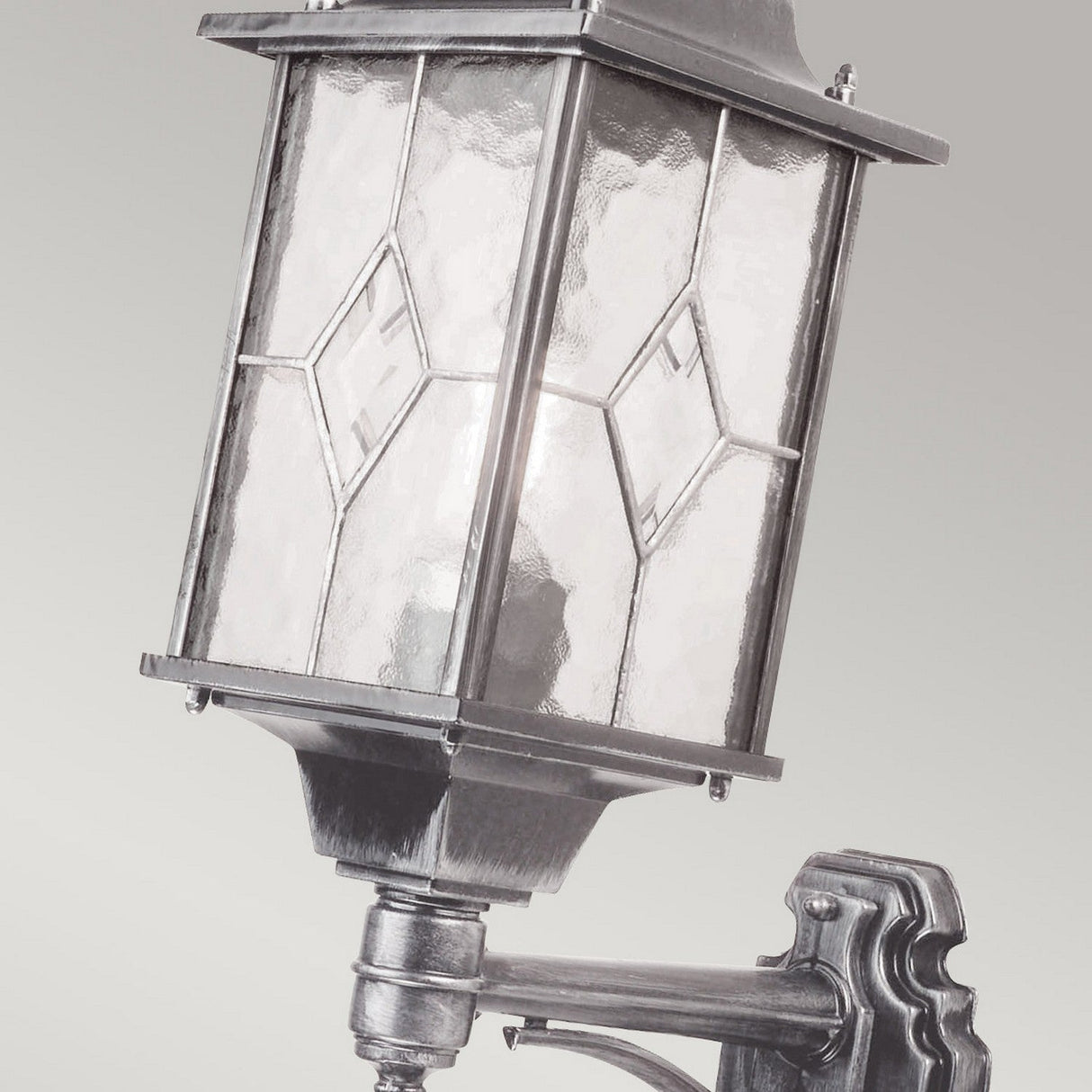 The Wexford 1 Light Outdoor Up Wall Light - Black/Silver features a vintage-style design with a metal frame and clear leaded glass panels. Its geometric patterns echo the traditional lantern shape, creating an elegant touch against a simple, neutral background.