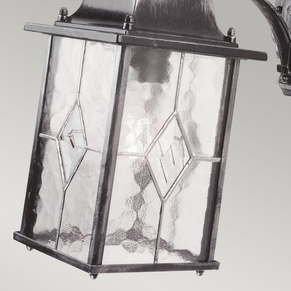 A close-up of the Wexford 1 Light Outdoor Down Wall Light showcases its vintage-style design with textured glass panels and diamond-shaped patterns. The weather-resistant fixture features a metal frame in a black/silver finish, providing illumination and casting a soft glow on the surroundings.