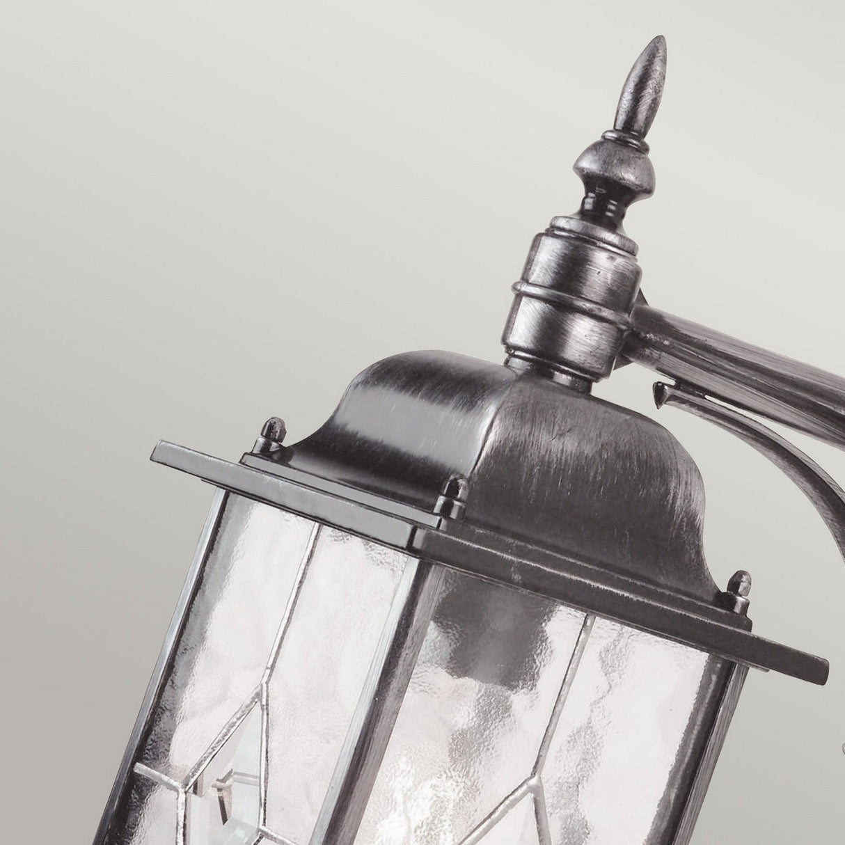 Close-up of the Wexford 1 Light Outdoor Down Wall Light in Black/Silver, featuring a metal frame and clear glass panels with decorative detailing on the top. This weather-resistant lantern stands out against a plain, light gray background.
