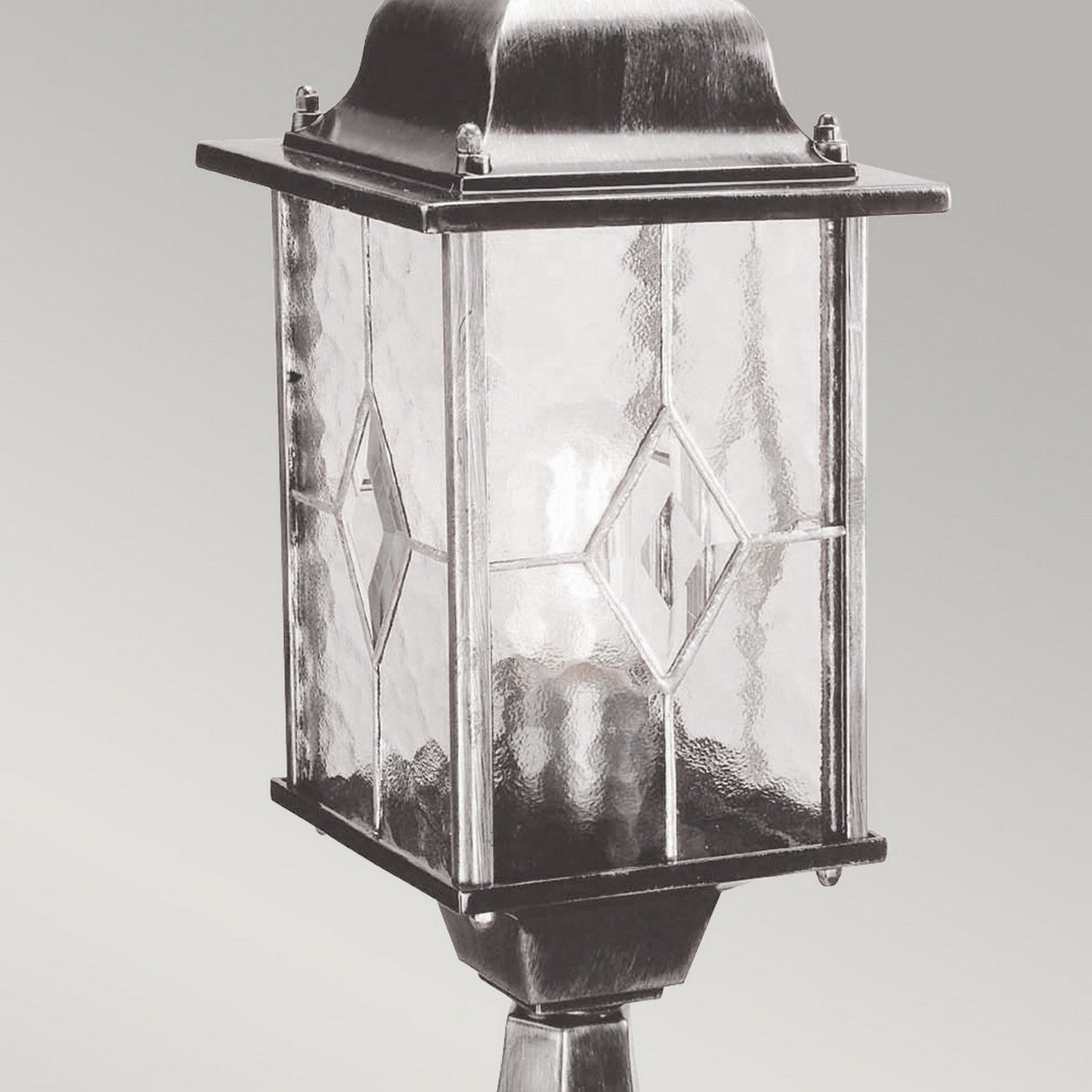 The Wexford 1 Light Pedestal Light, with its vintage-style design, features a metal frame and textured glass panels arranged in a diamond pattern. It stands elegantly against a neutral gray background.