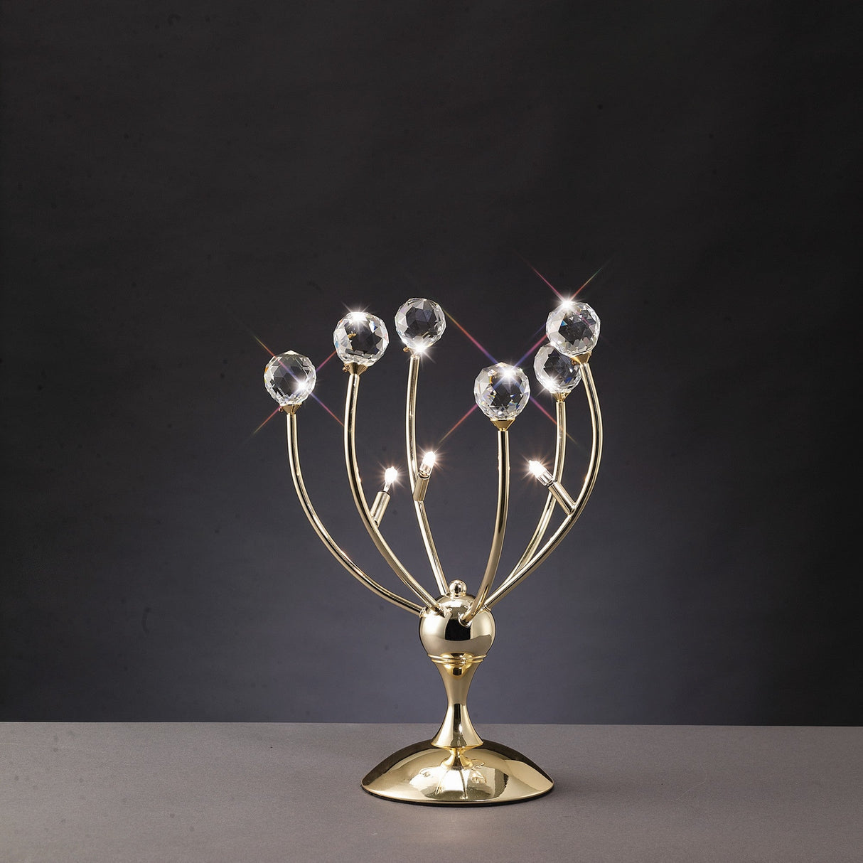 The Xeena 3 Light Crystal Table Lamp in French Gold features a modern, curved design with three branches topped by large faceted crystal spheres, displayed on a gray surface against a dark background.