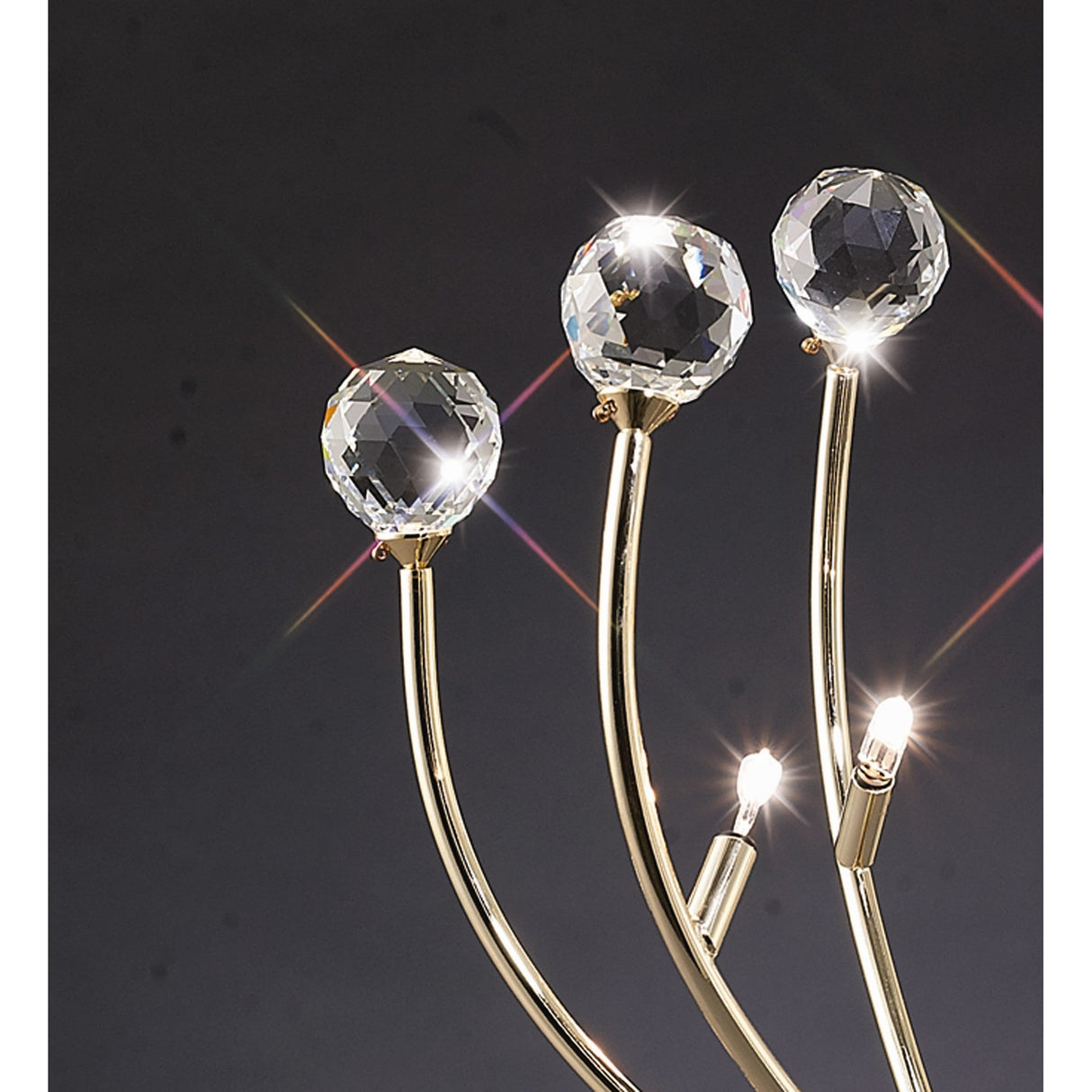 The Xeena 3 Light Crystal Table Lamp in French Gold features four curved metal arms holding three faceted crystal spheres and two LED lights, casting star-like sparkles against a dark background for a modern look.
