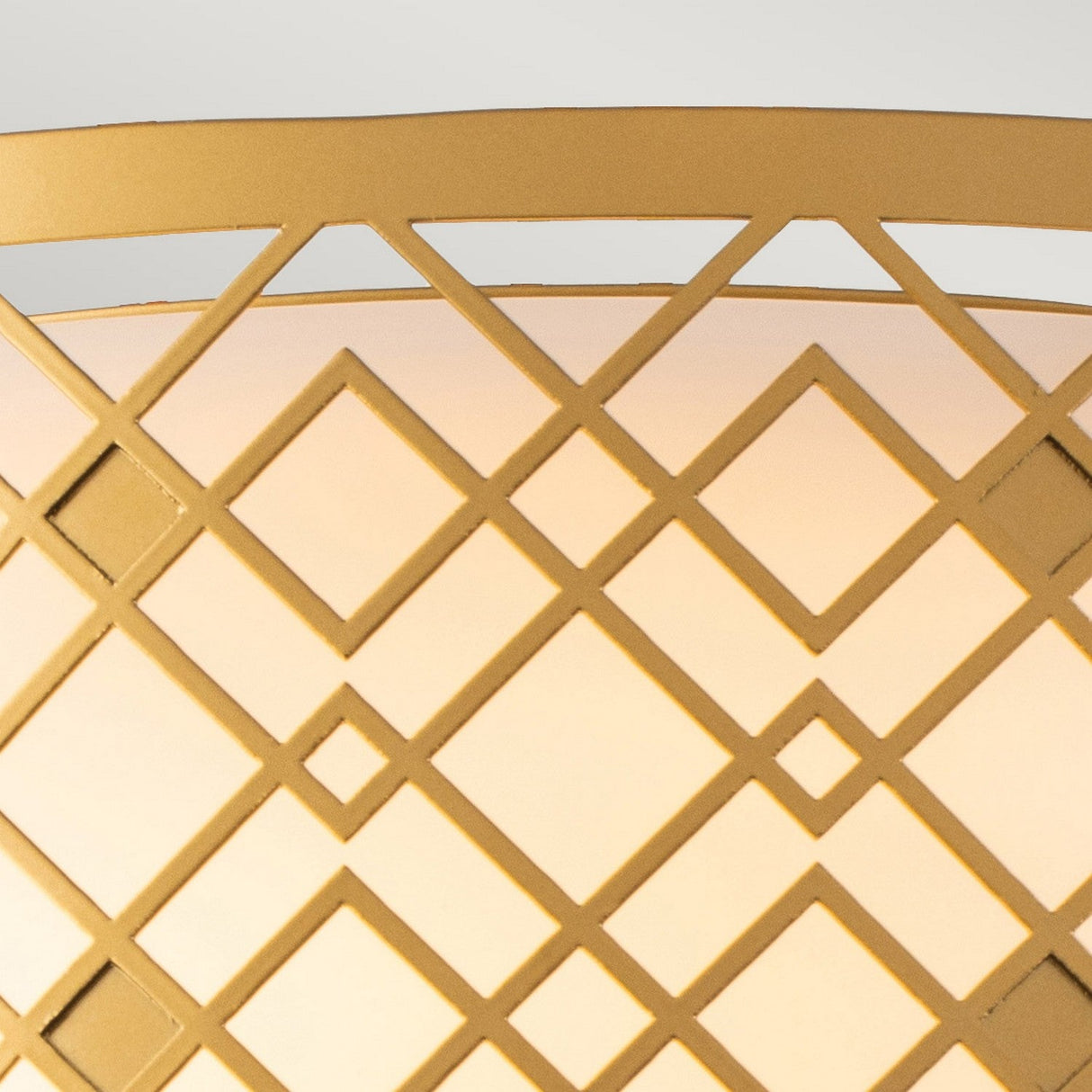 Close-up of the Ziggy 2 Light Medium Flush in lacquered gold, showcasing a geometric lamp shade with an intricate diamond and crisscross pattern. It emits a warm glow that highlights its sleek framework against a soft background, providing a contemporary look to elevate any space.