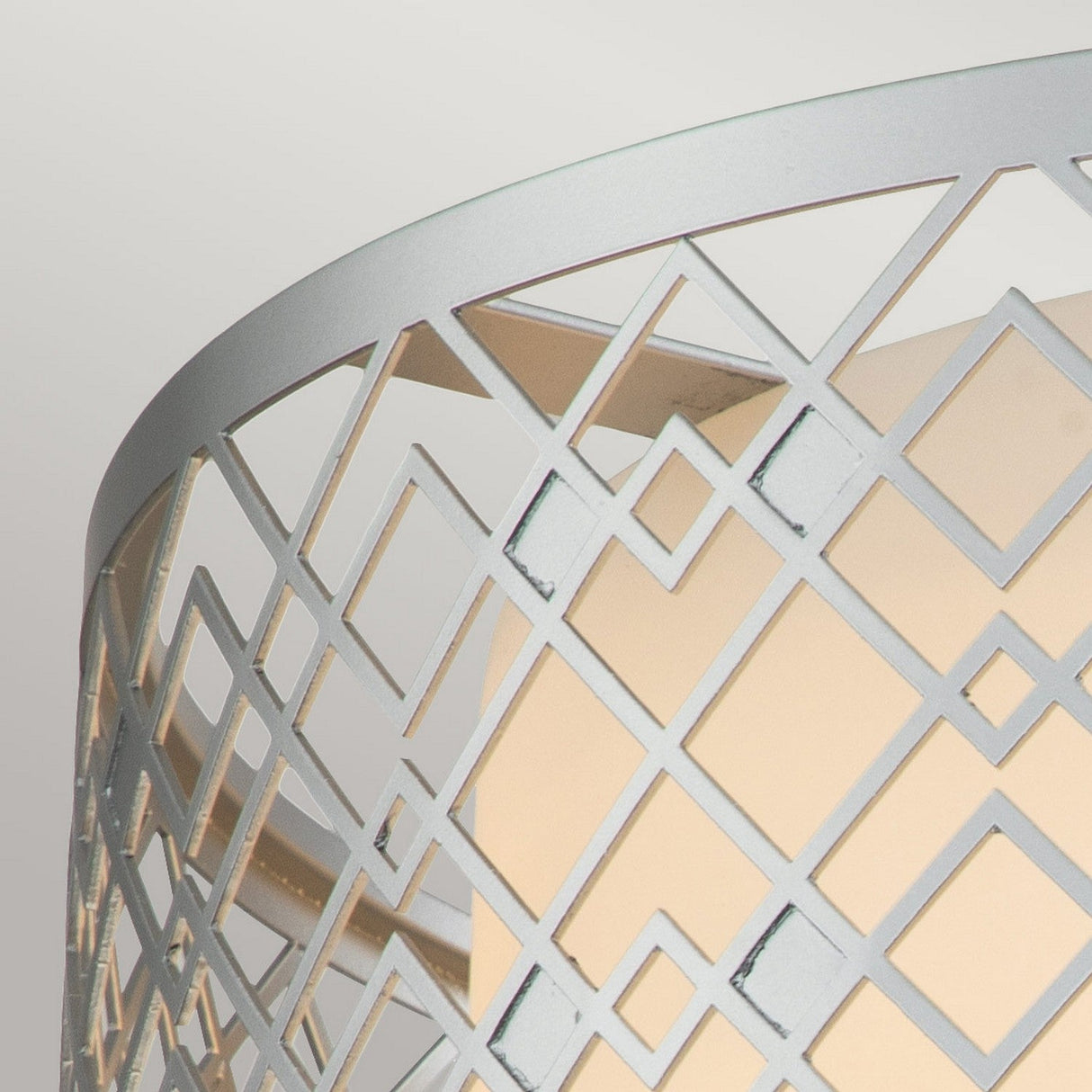 A close-up of the Ziggy 2 Light Medium Flush in Lacquered Silver reveals an intricate geometric lattice design with diamond shapes. The metallic lampshade emits a soft, warm glow from within, reminiscent of a modern flush light, against a pale background.