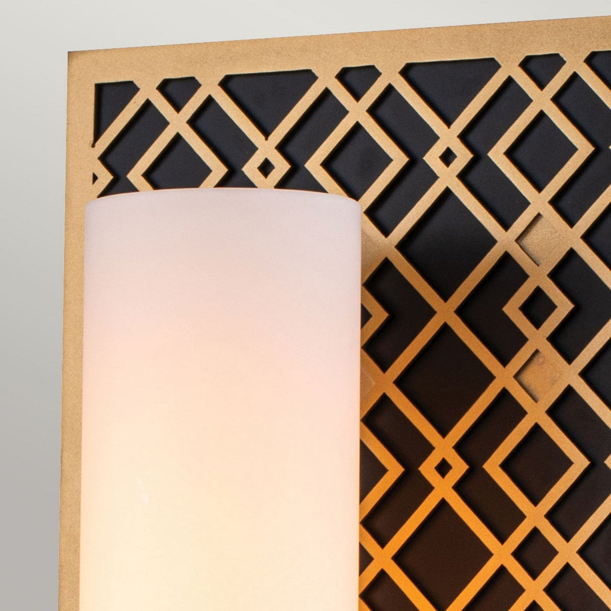 A close-up view of the Ziggy 1 Light Wall light - Lacquered Gold reveals its cylindrical white shade, mounted on a textured golden metal panel. This panel features a black background with a diamond-shaped cutout pattern, highlighting its modern design. The light is turned on and boasts an IP44 rating.