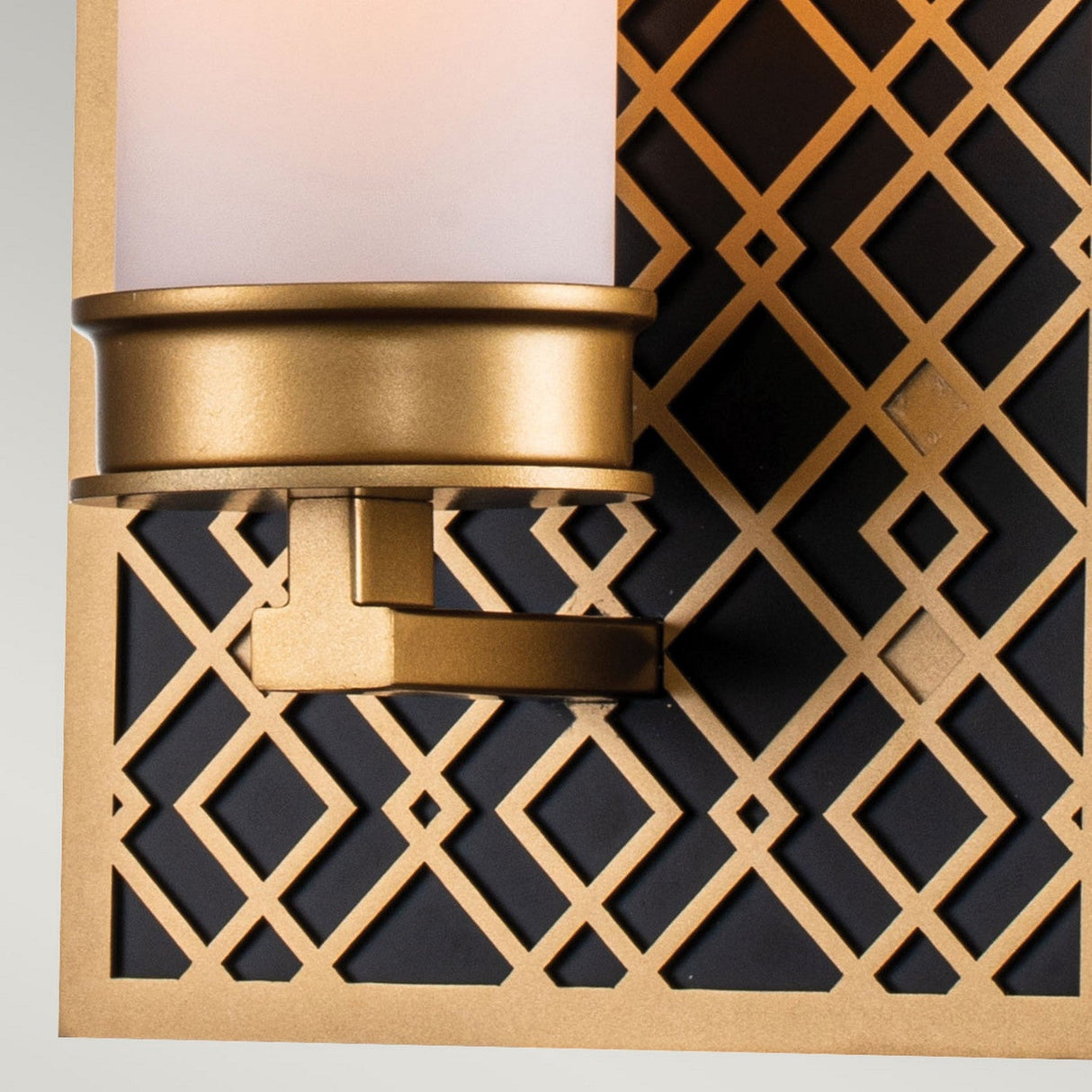 Close-up of the Ziggy 1 Light Wall Light in Lacquered Gold, featuring a decorative design with a frosted glass shade and gold metal elements. This modern wall sconce perfectly complements the background's intricate geometric lattice and diamond patterns.
