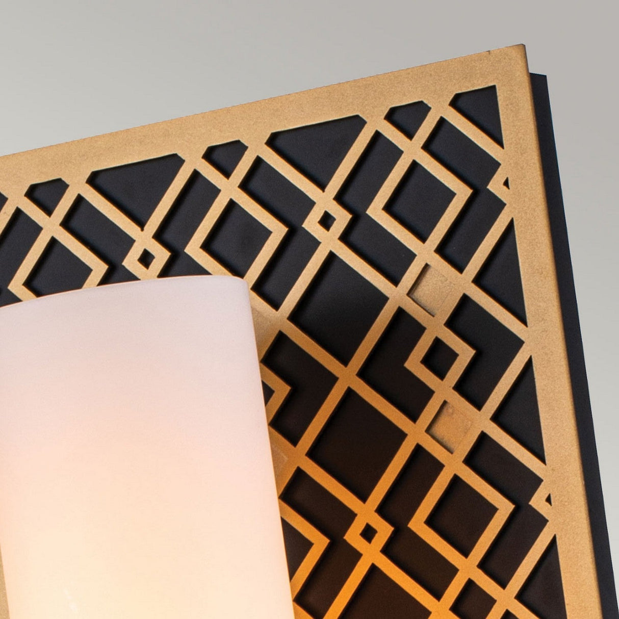 Close-up of the Ziggy 1 Light Wall Light - Lacquered Gold, a decorative fixture featuring a geometric lattice pattern in black and gold with a frosted glass shade. Its modern design enhances any setting against the simple light gray background.